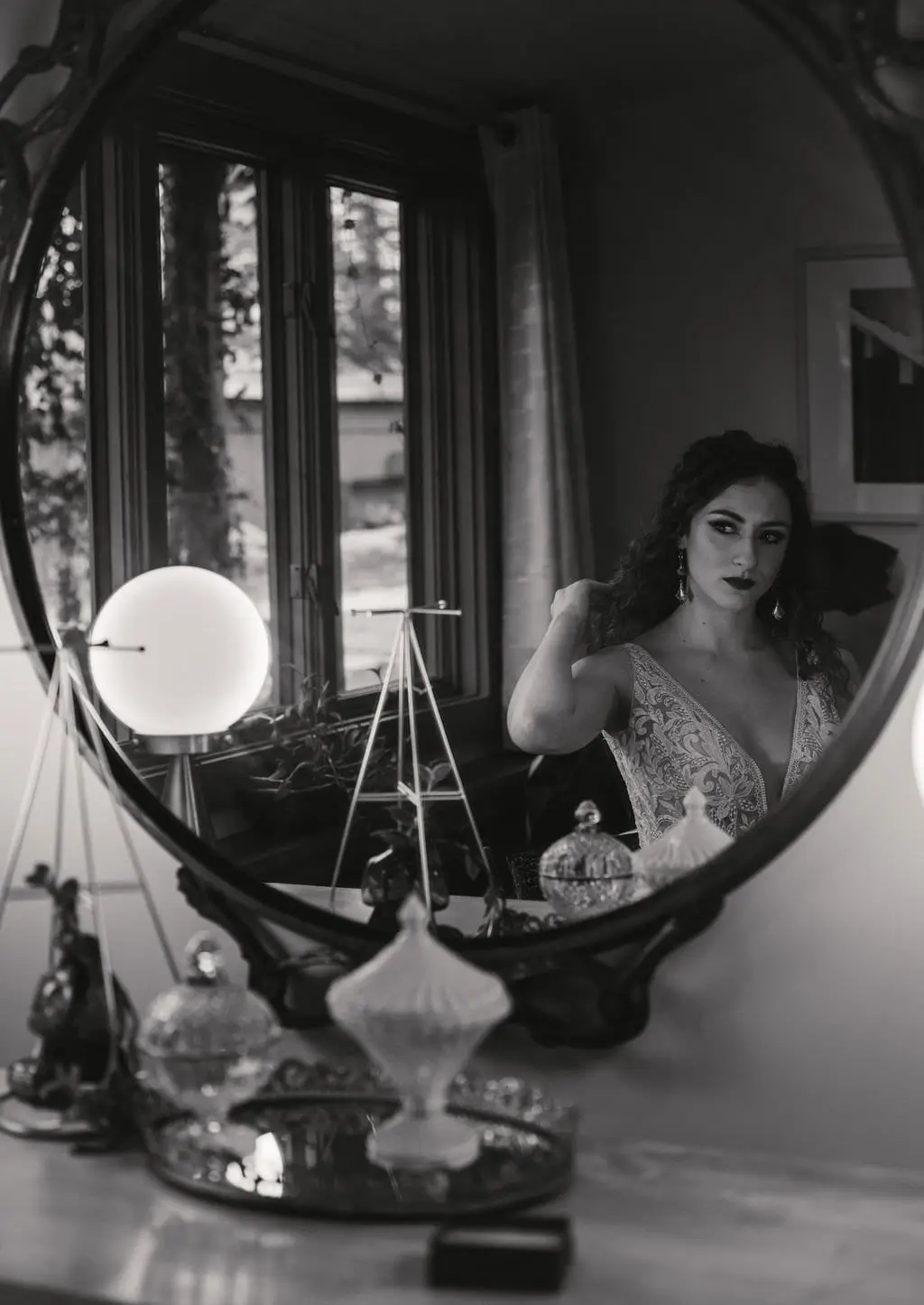 Black and white reflection of the bride in a round vanity mirror among perfume bottles during getting ready