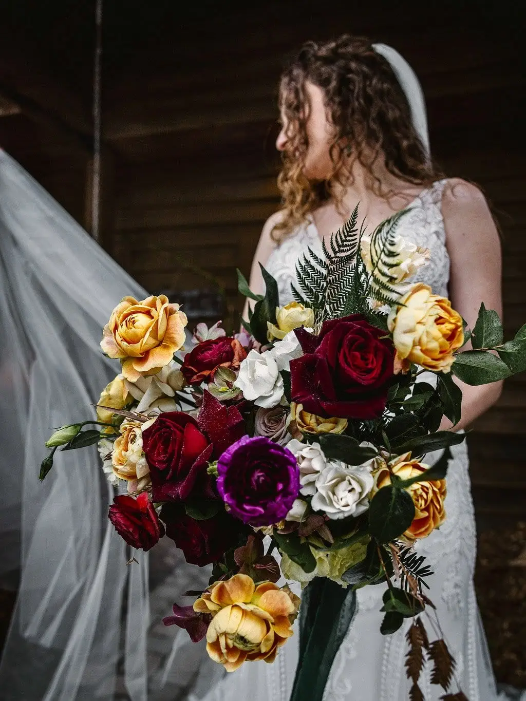 Bride looks down at her red, gold and purple bouquet as her veil drifts beside her