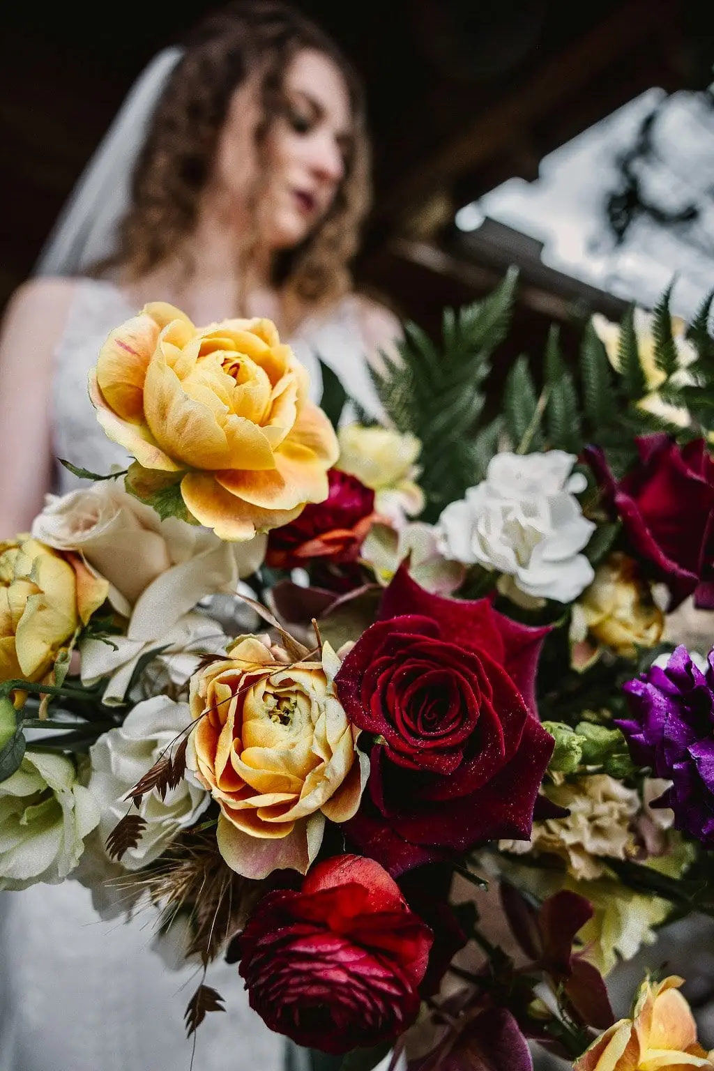 Soft-focus bride behind a bouquet of golden roses, red ranunculus and white blooms