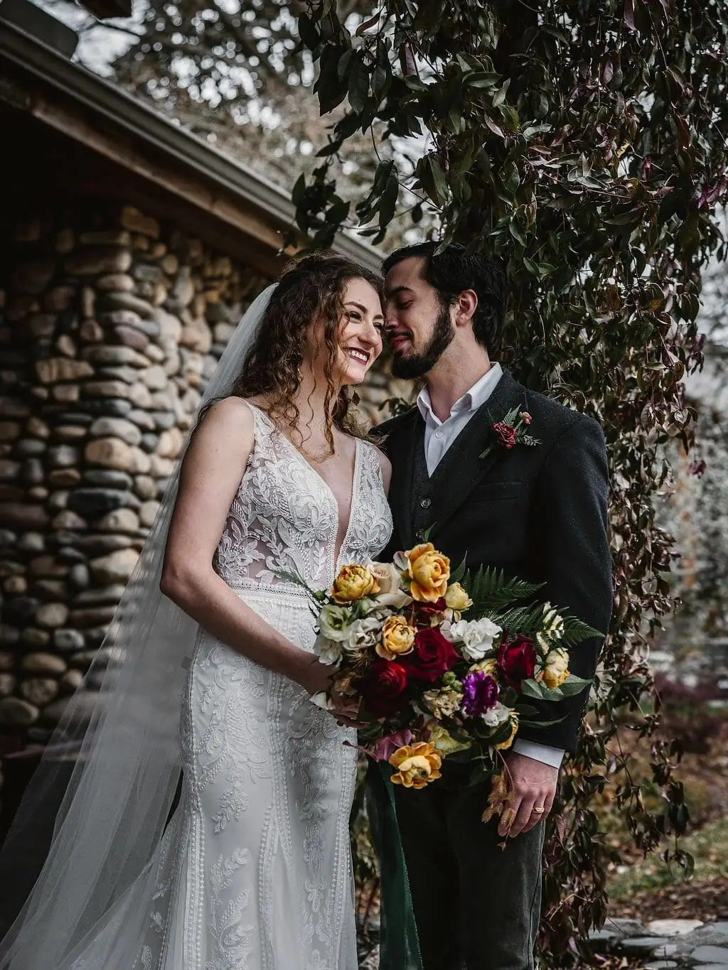 A couple smiling together with a bouquet of red and yellow roses outside Haiku I Do