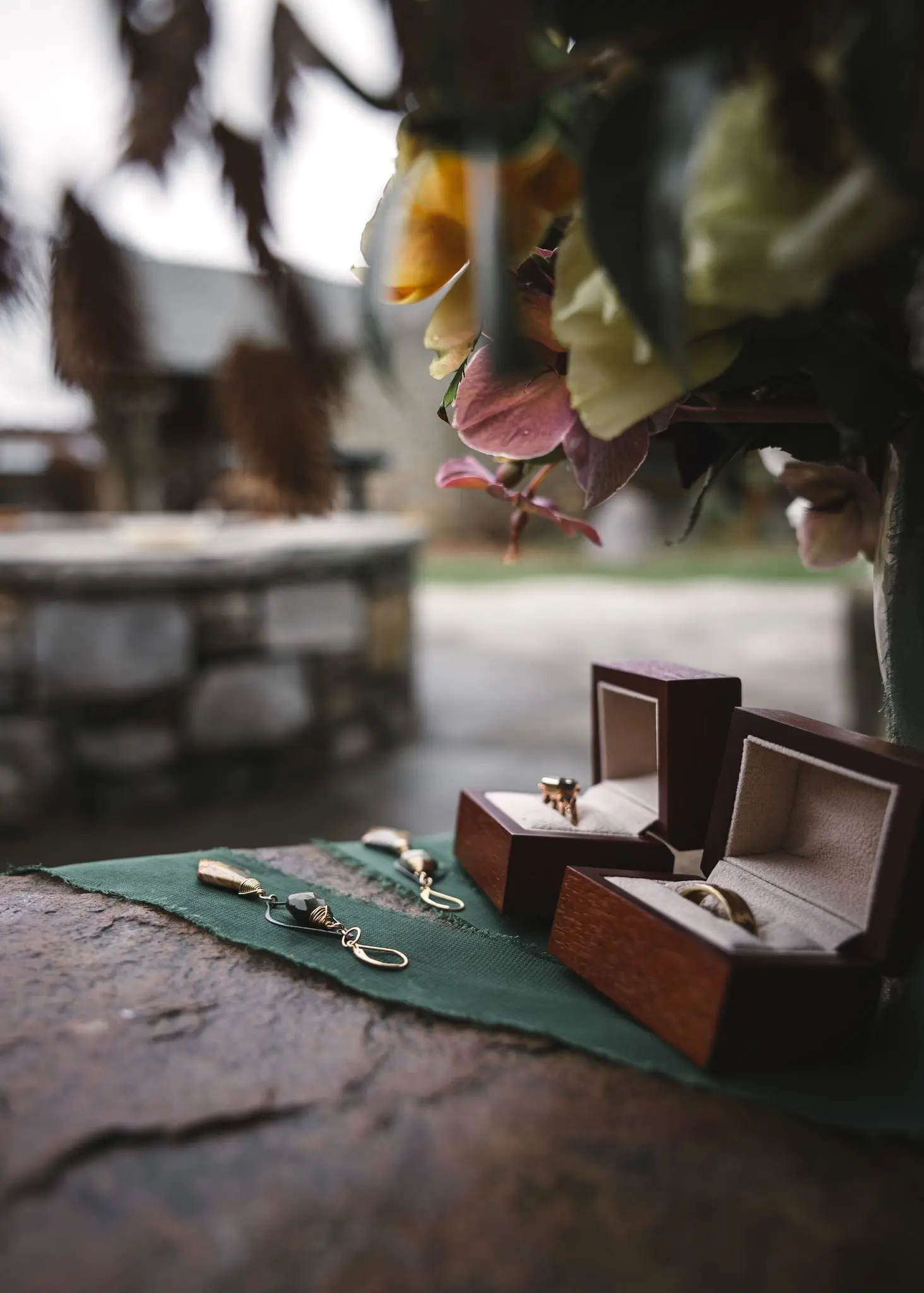 Wedding rings in wooden boxes and earrings on a green cloth beside a floral arrangement and stone fire pit