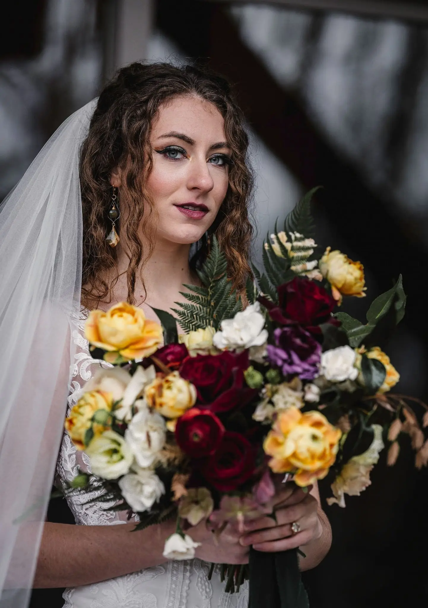 Bride with curled hair and a long veil holds an autumn-toned bouquet and looks into the camera