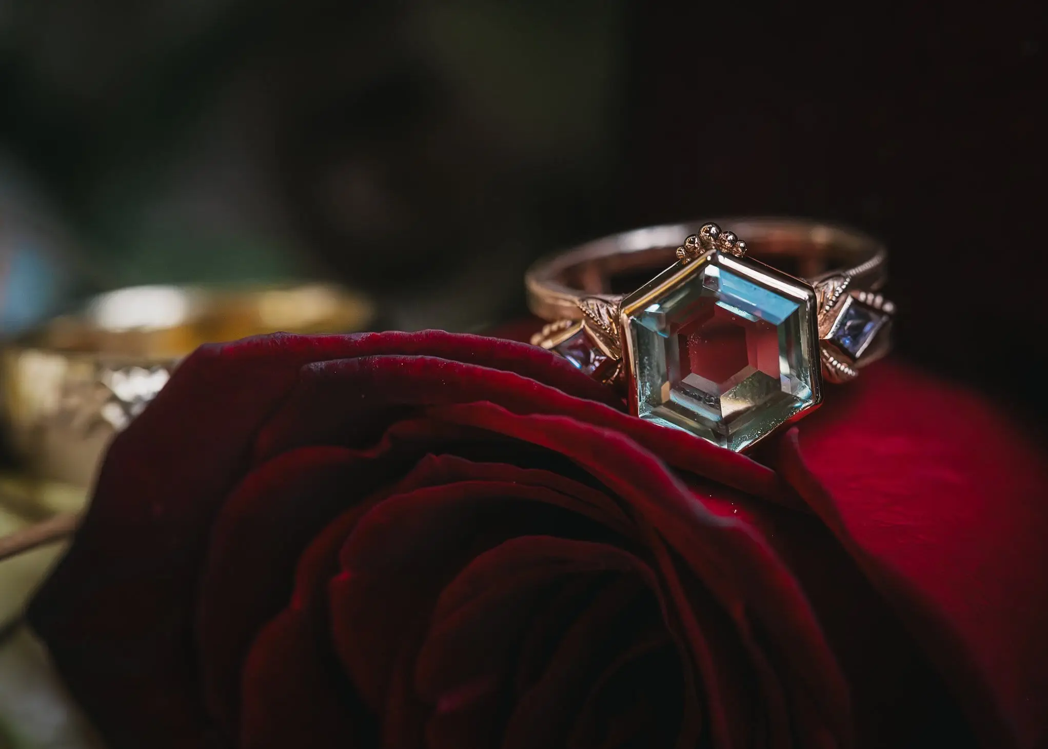 A silver ring with a large, hexagon-shaped, multi-colored gemstone resting on a dark red rose.