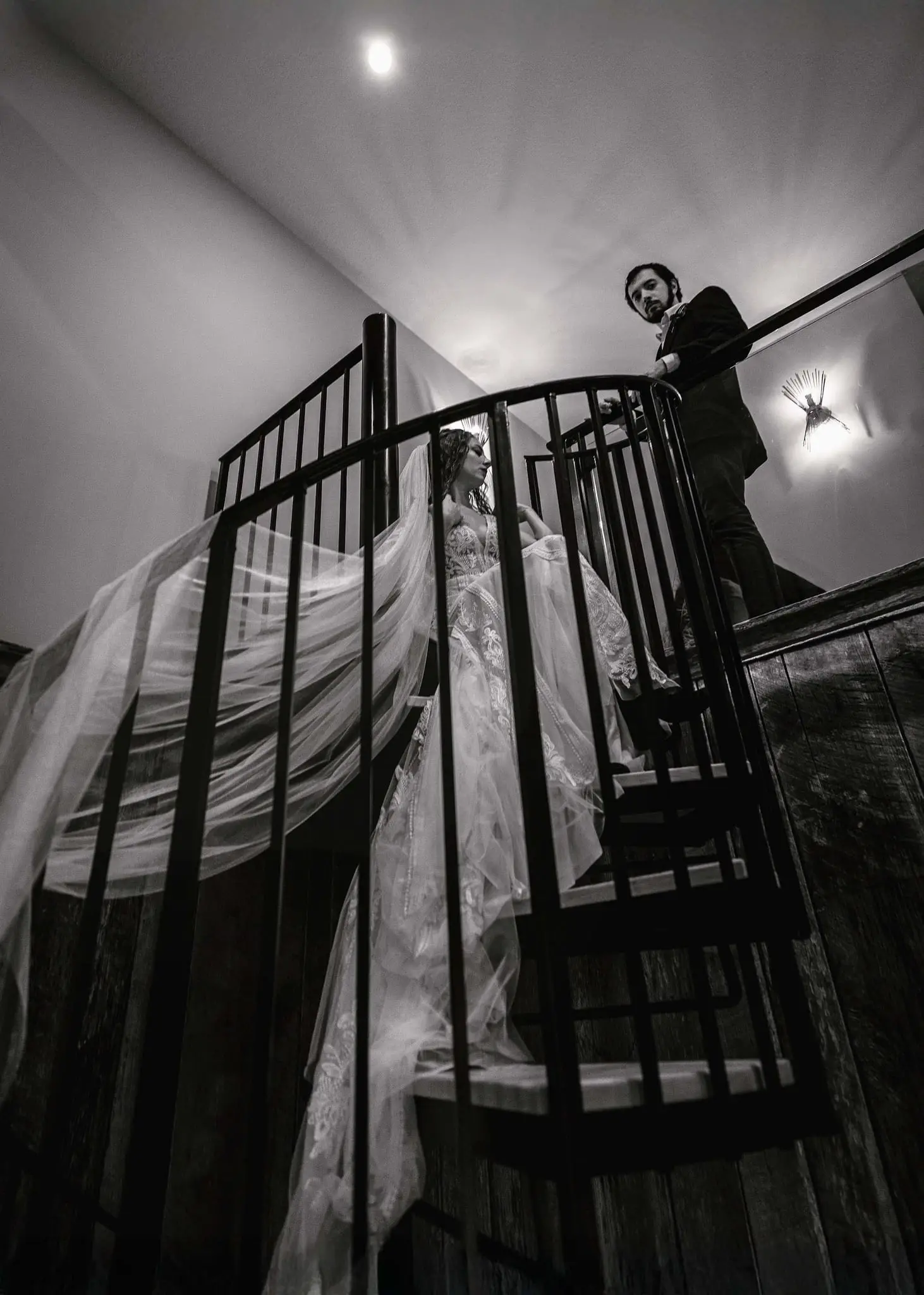 Dramatic black and white shot of the bride's long veil flowing down a spiral staircase as the groom looks on