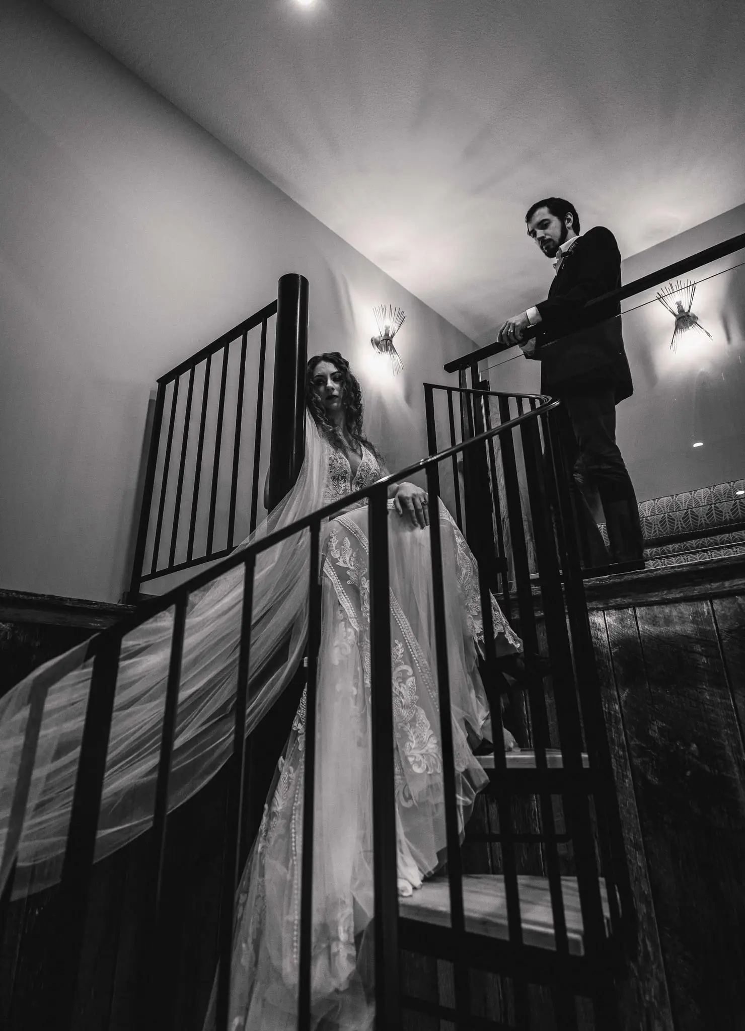 Black and white view up a spiral staircase as the bride descends in her gown while the groom watches from above