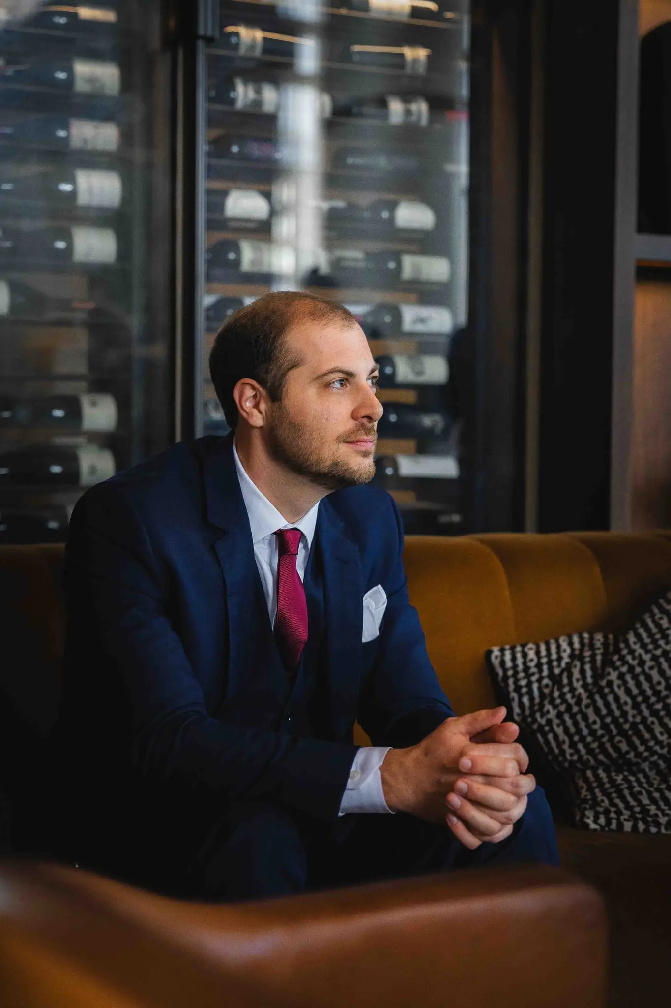 Groom in a navy suit and burgundy tie seated on a leather sofa beside a glass wine wall