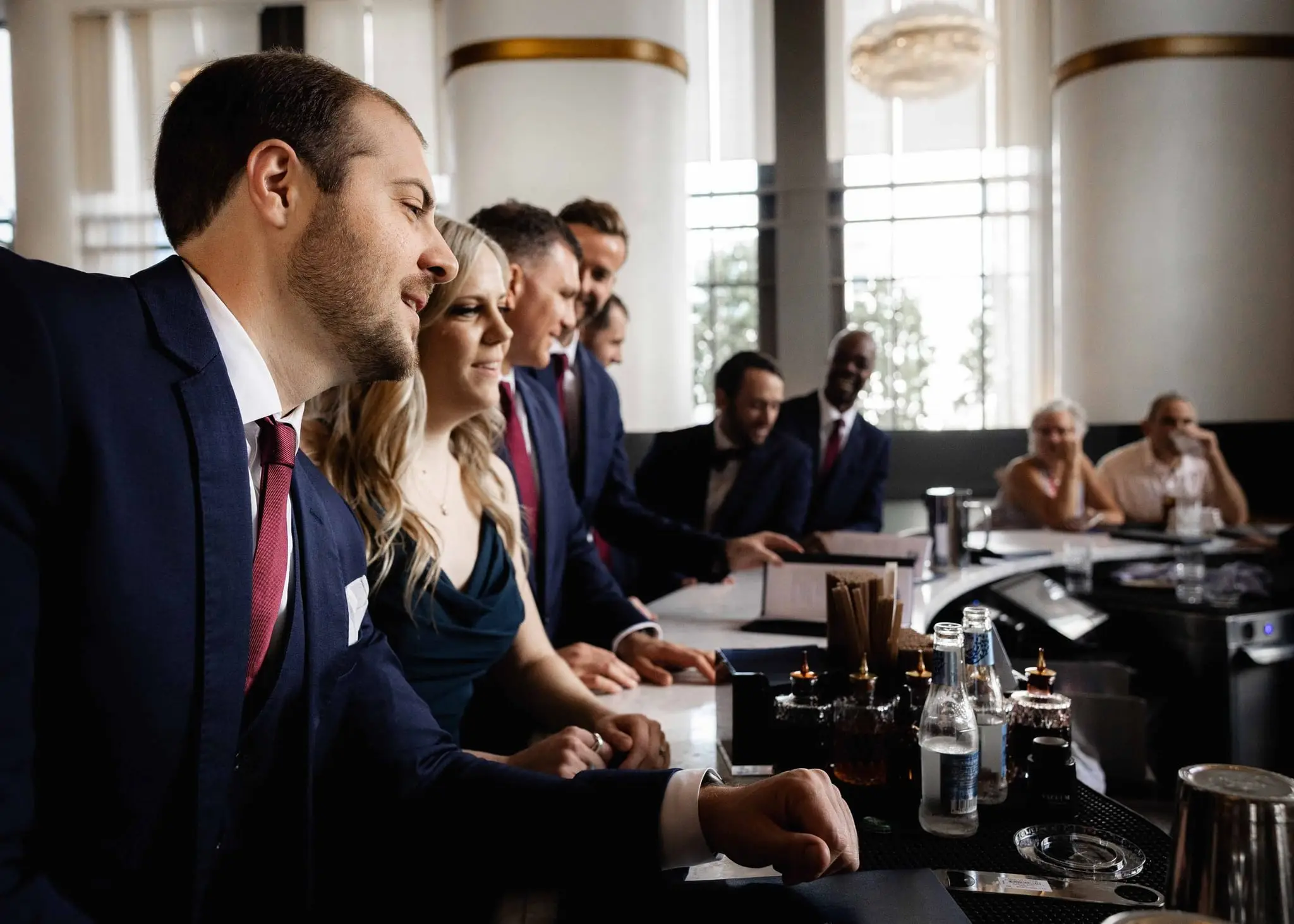Groom, groomsmen and a bridesmaid laughing together along a curved hotel bar