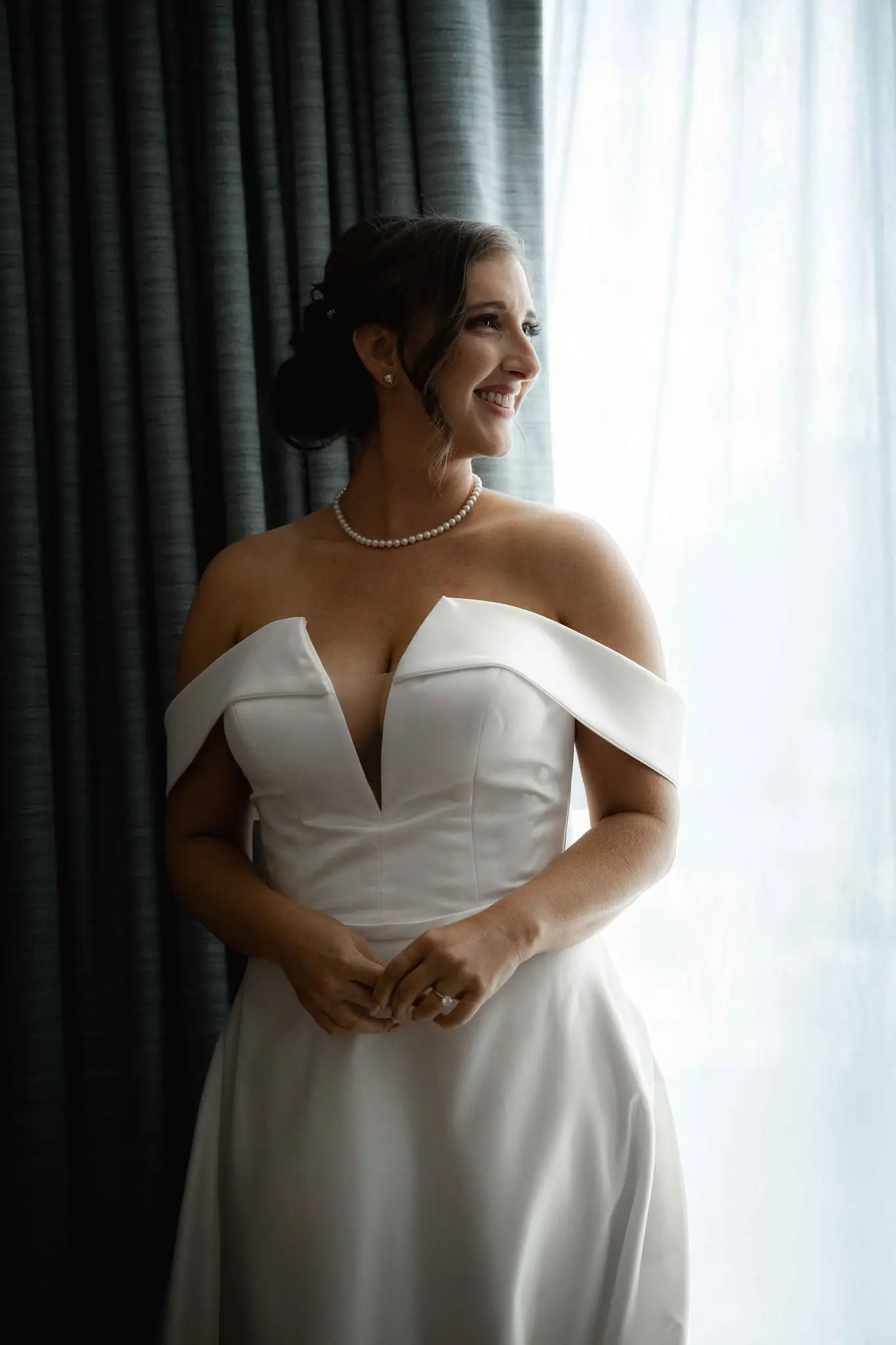 Ashley in her off-shoulder wedding dress smiling by a bright window in her suite at the Conrad Nashville