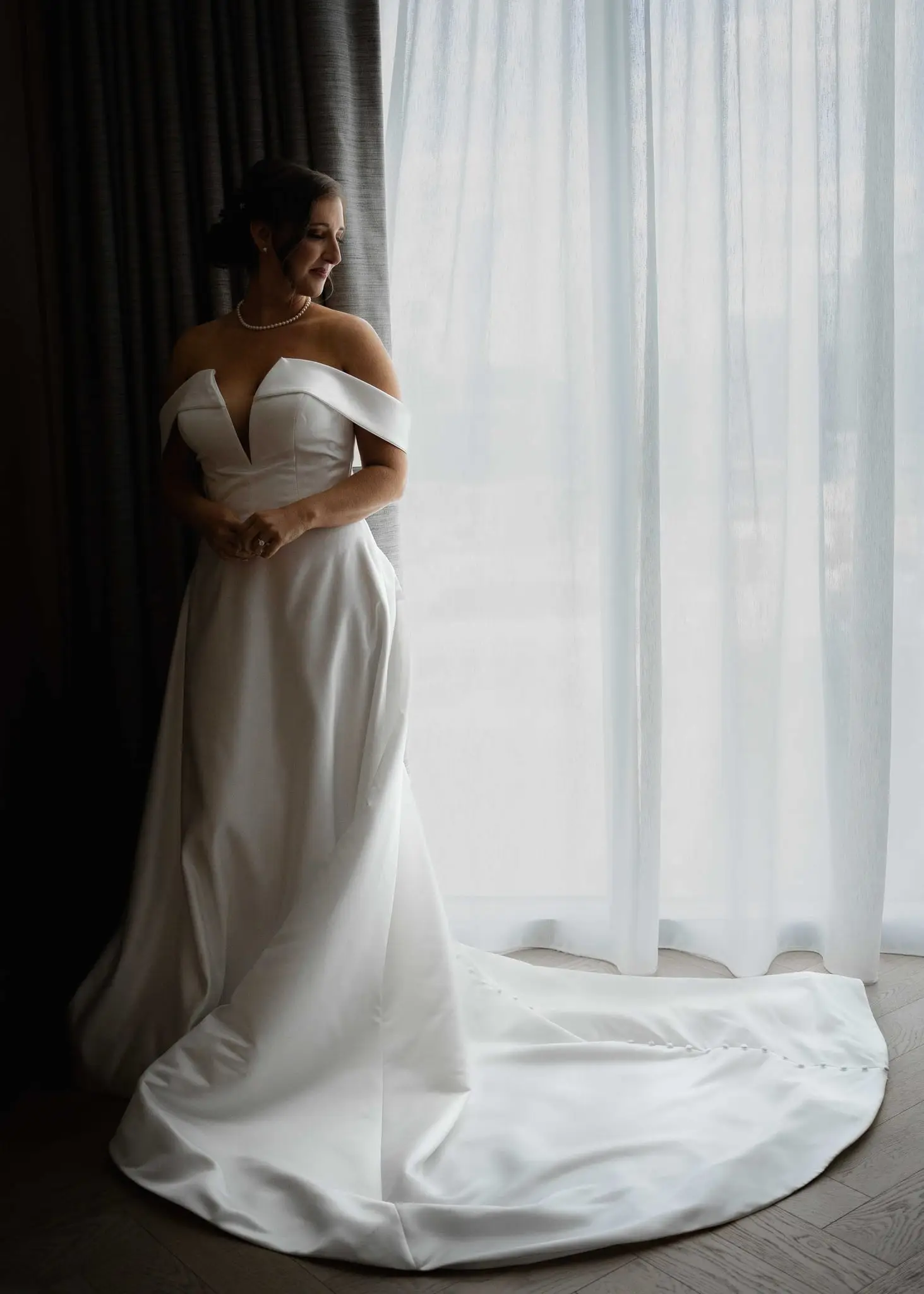 Full-length portrait of the bride by sheer curtains, her gown train spread across the floor