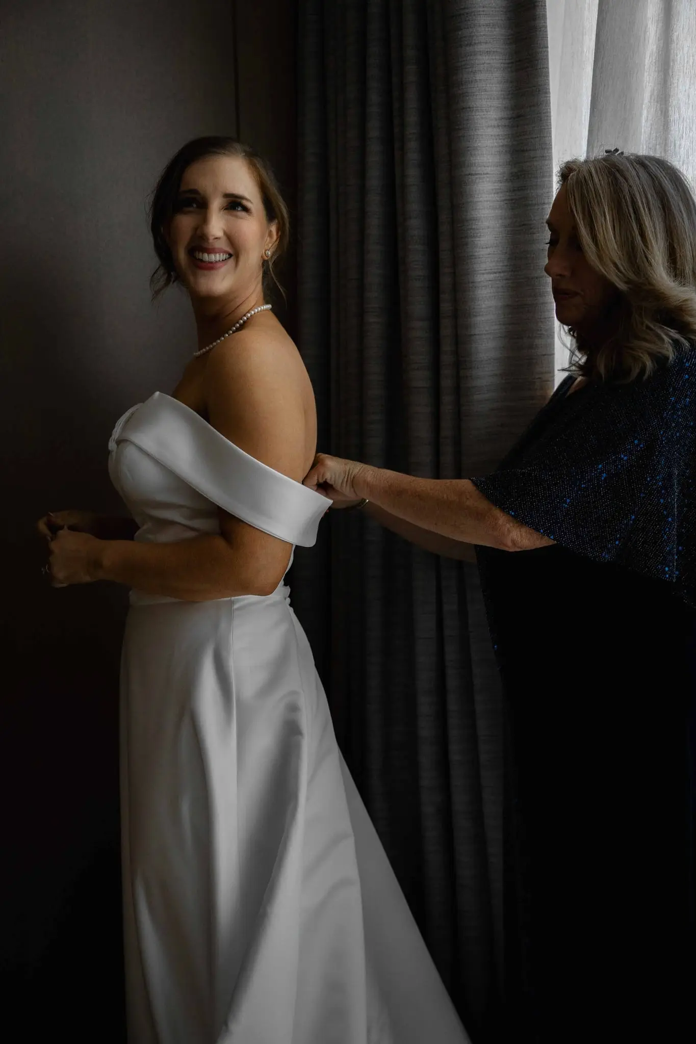 Bride smiling over her shoulder as an older woman fastens the back of her dress
