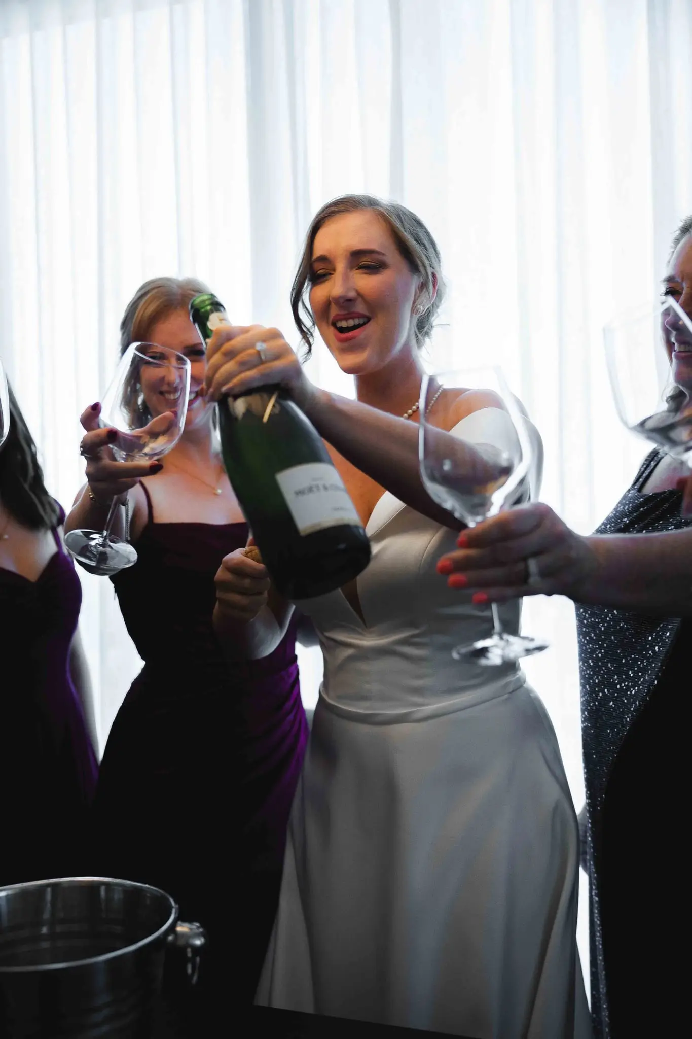 Bride opening a champagne bottle and laughing with bridesmaids holding glasses