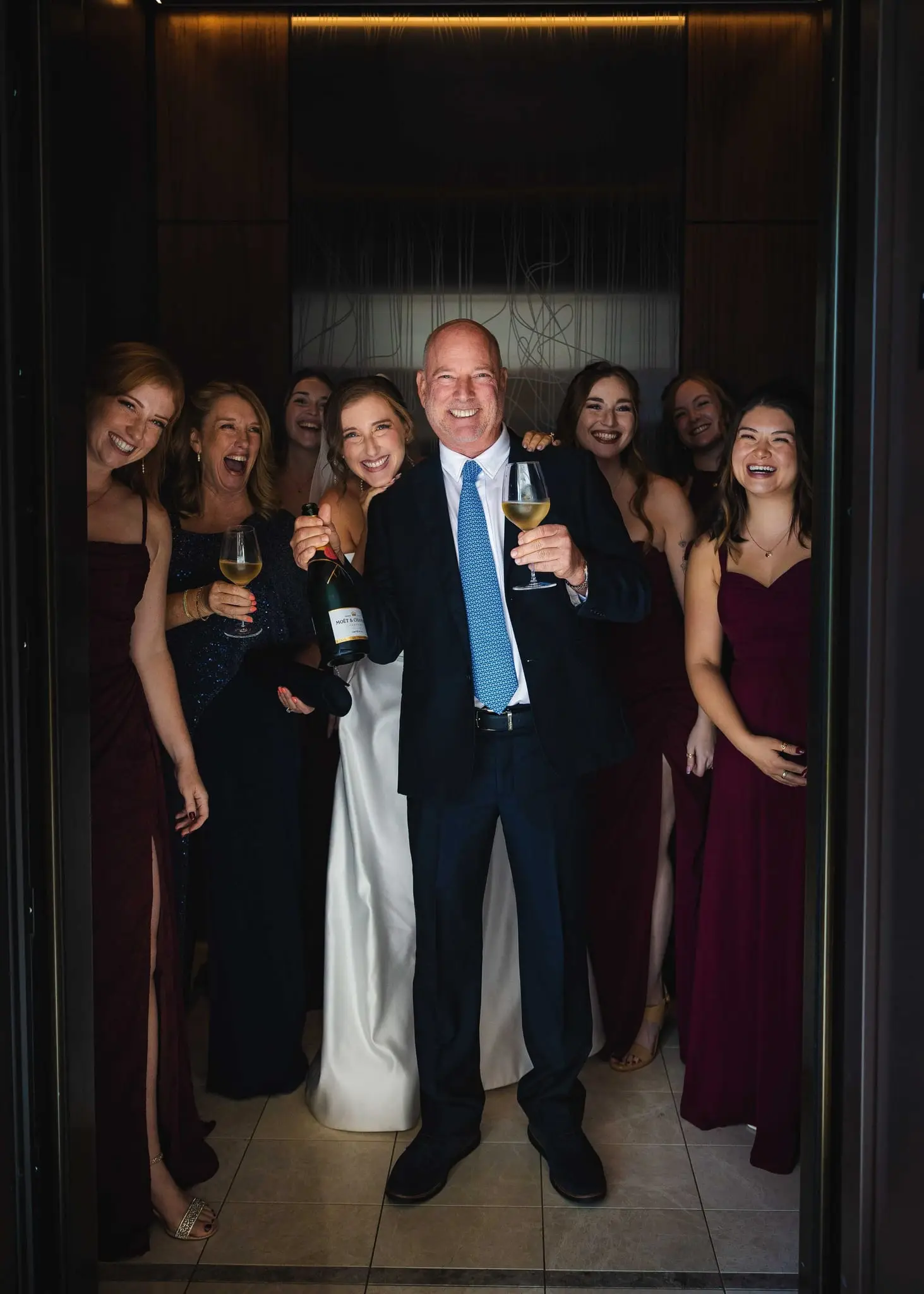 Bridesmaids and a man in a blue tie crowd around the bride in a doorway, glasses raised