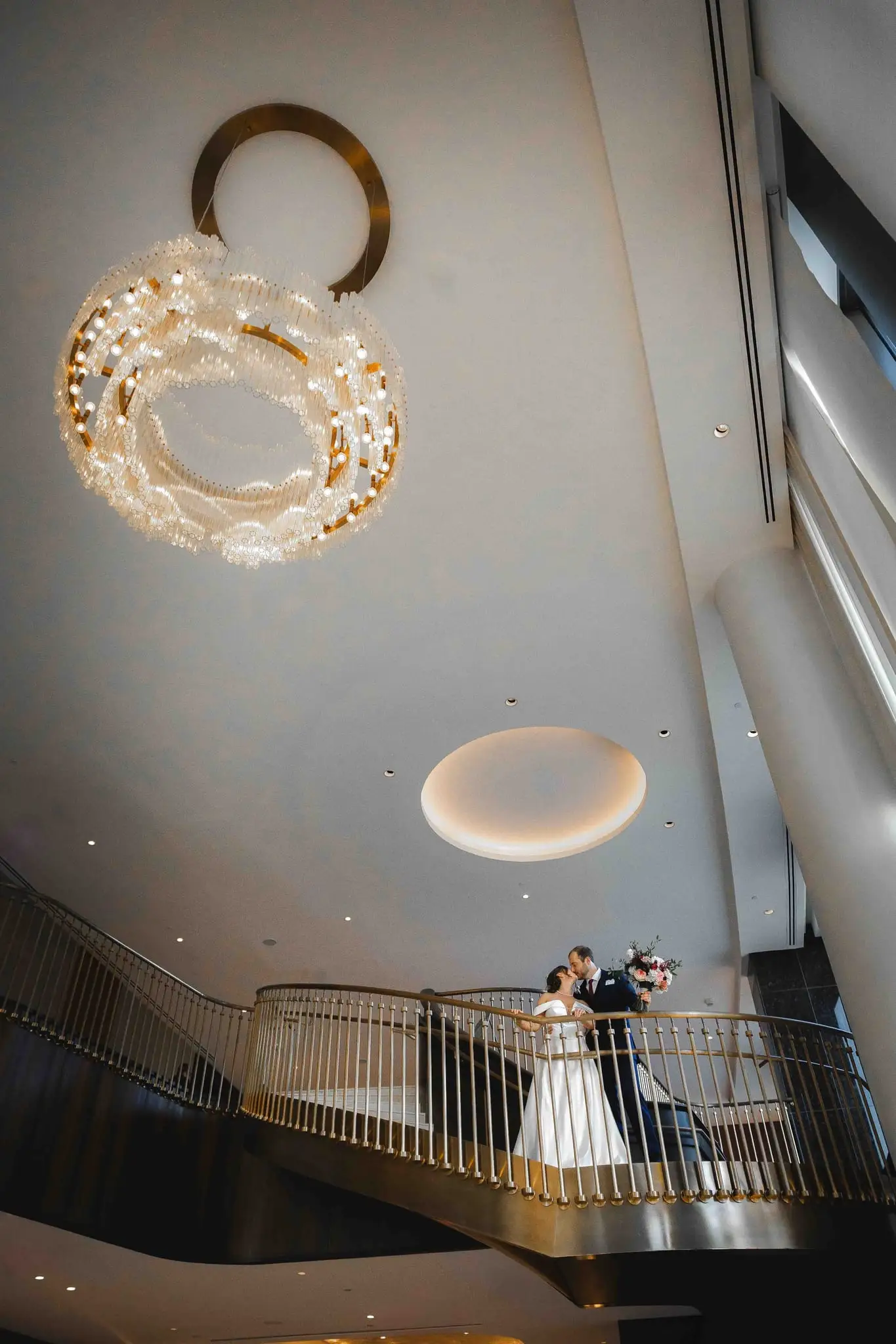 Ashley and Alex wedding at Conrad Nashville, Nashville TN — the couple on a gold-railed balcony seen from far below, a chandelier overhead