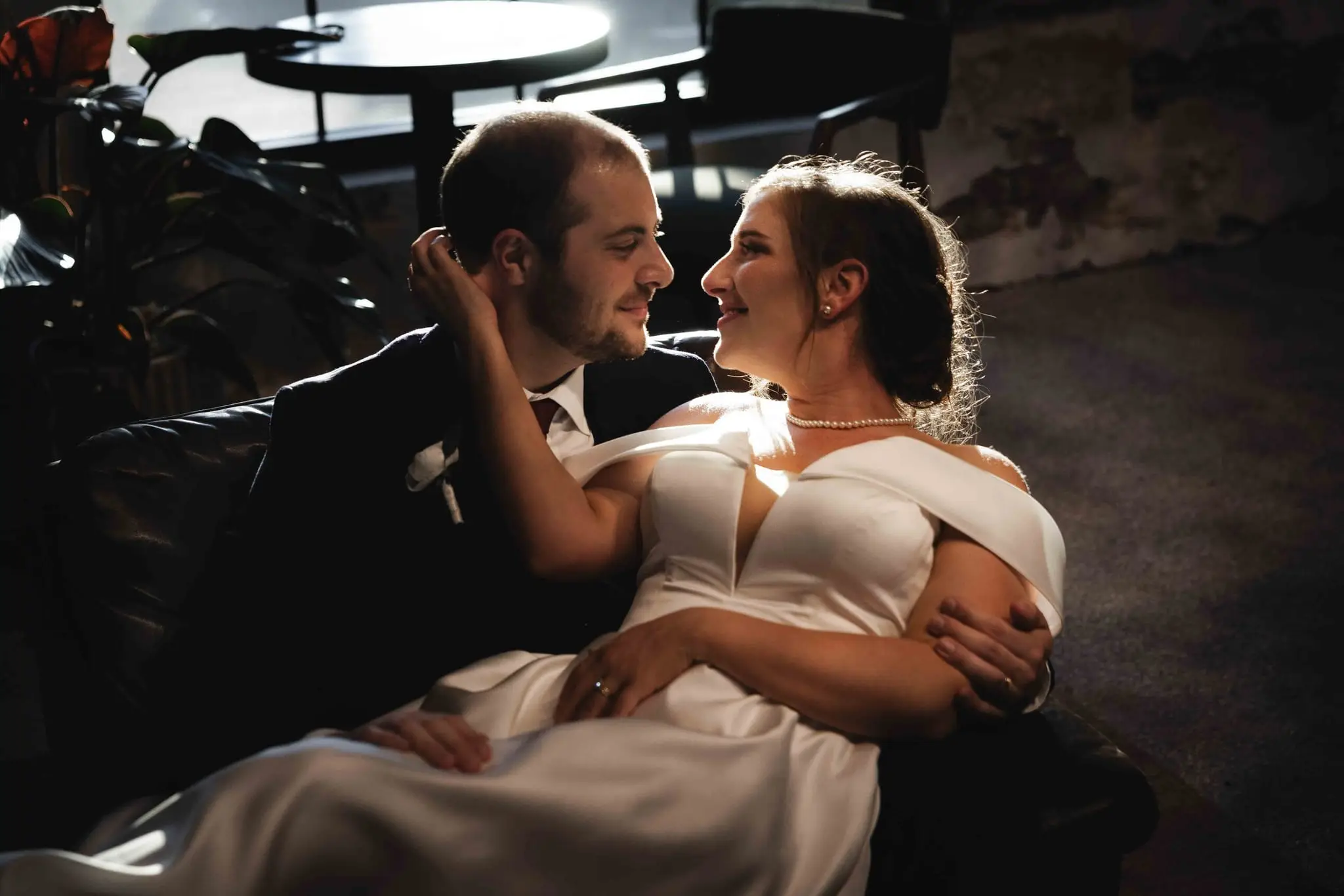 Groom leaning in toward the bride on the sofa as they smile, plants and a cafe table behind