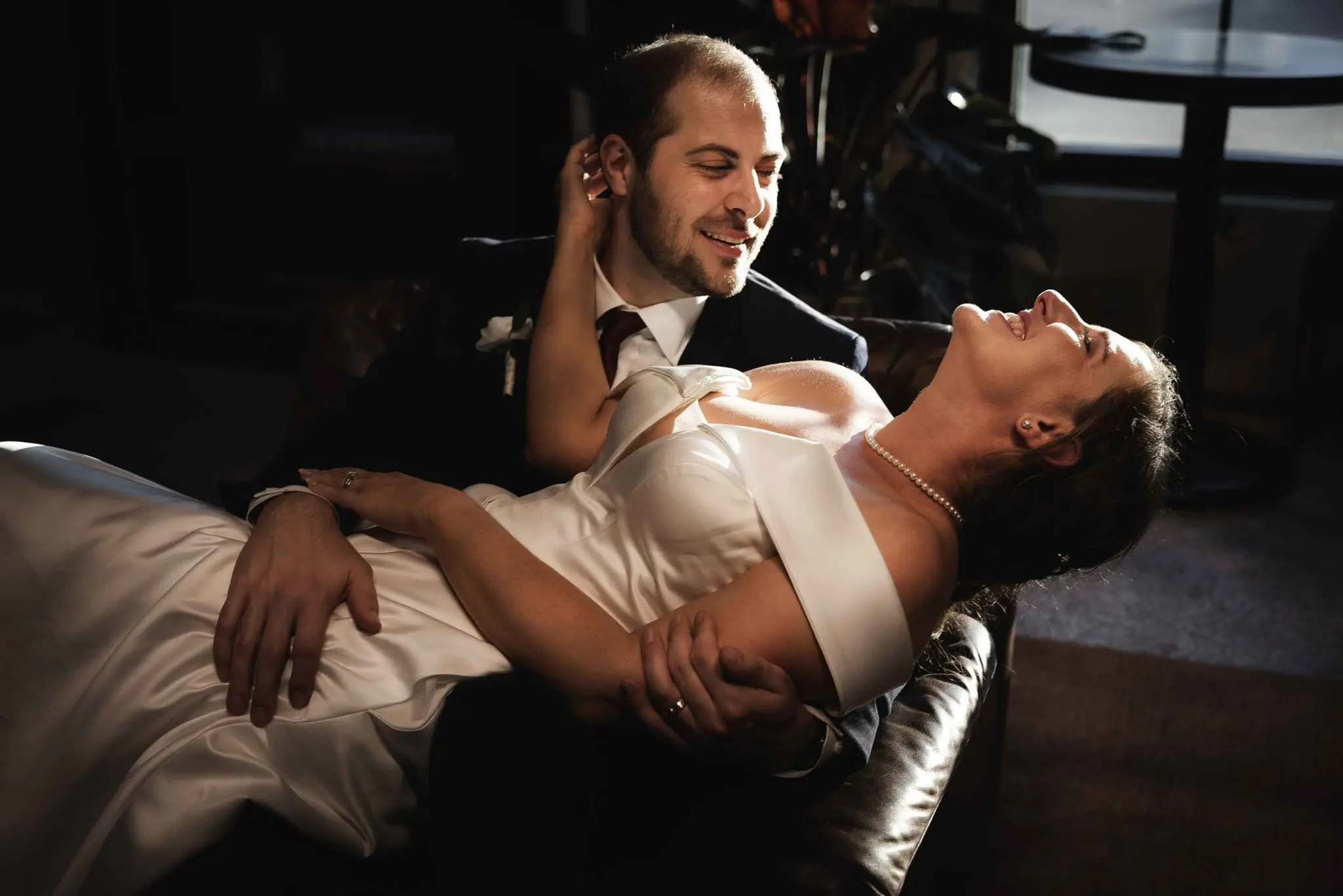 Groom smiling down at the bride as she lies back across his lap, pearls catching the light