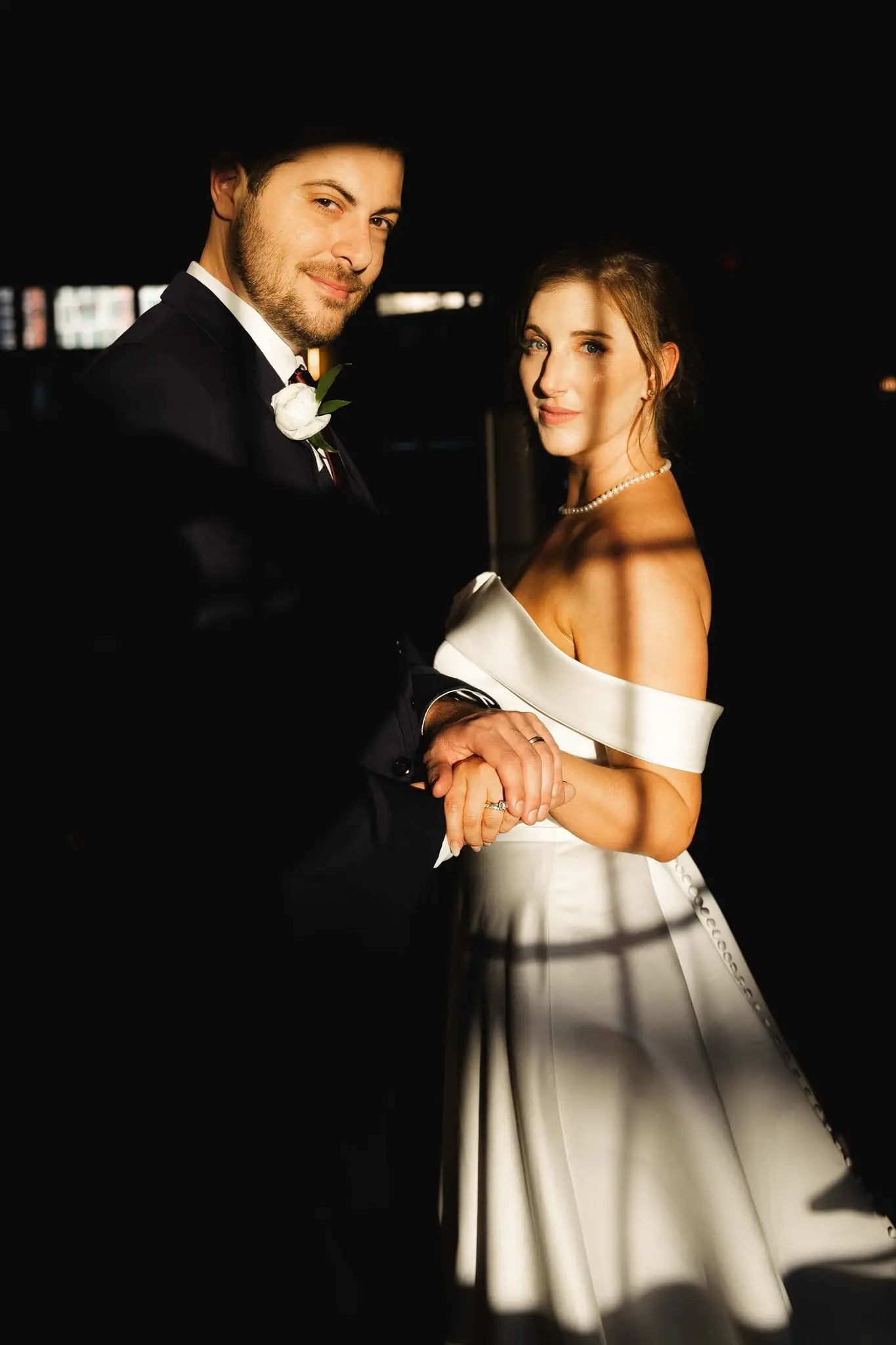 Couple standing close with clasped hands as striped window light falls across the bride's dress