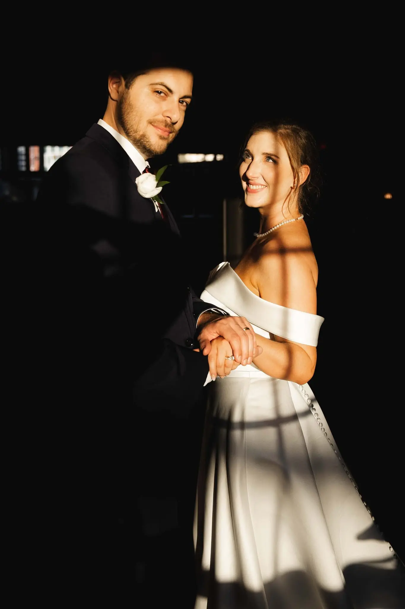 Ashley and Alex smiling at the camera in a pool of window light, hands clasped