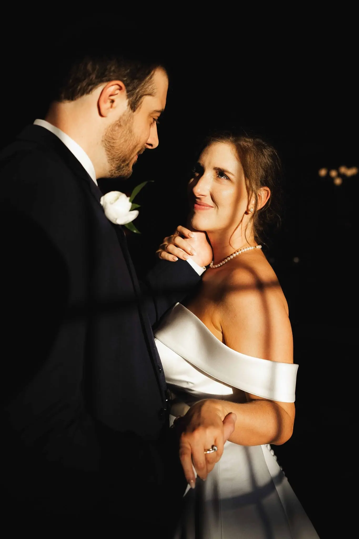 Groom gazing at the bride as she smiles back, shadows across her shoulder against a black backdrop