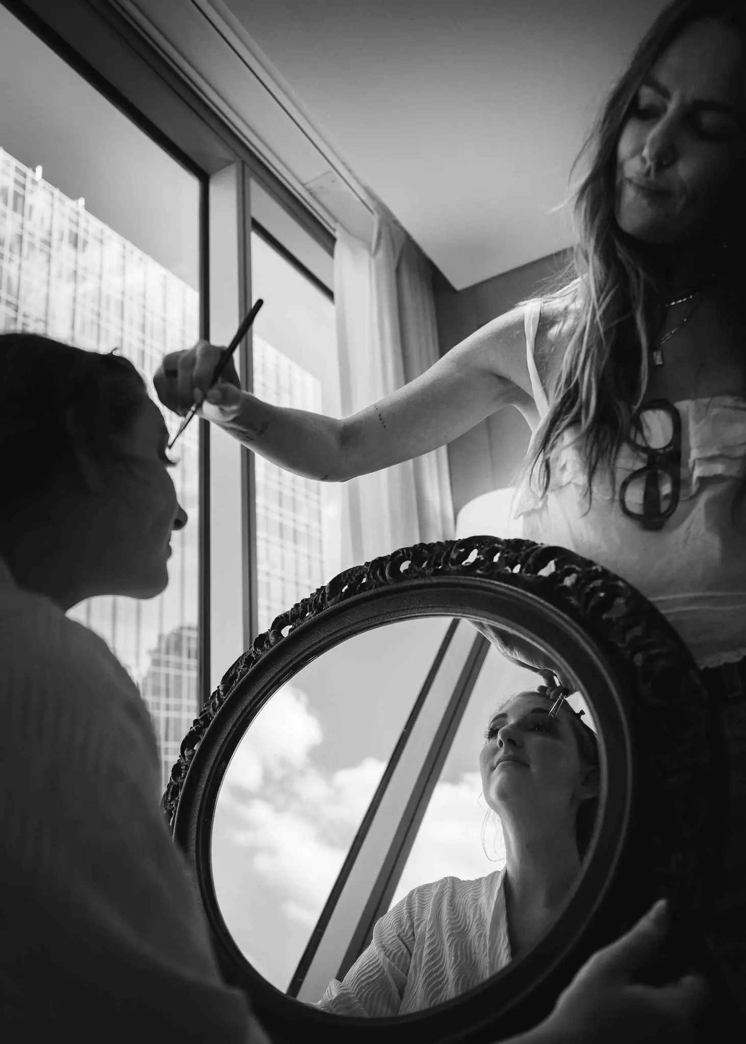 Black and white photo of the bride having makeup applied, her reflection in a round mirror