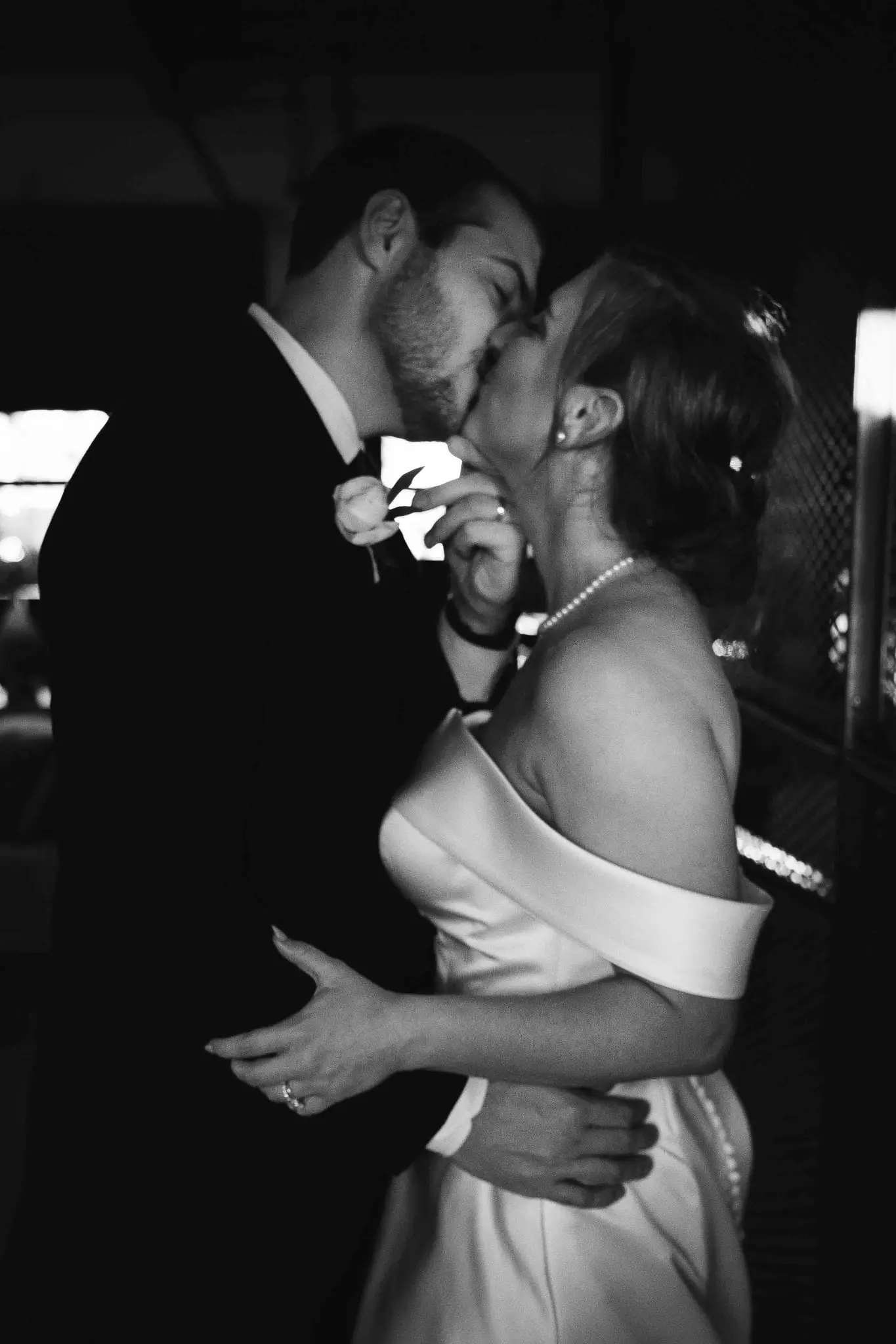 Black and white photo of a couple kissing, dressed in wedding attire, inside a dimly lit venue.