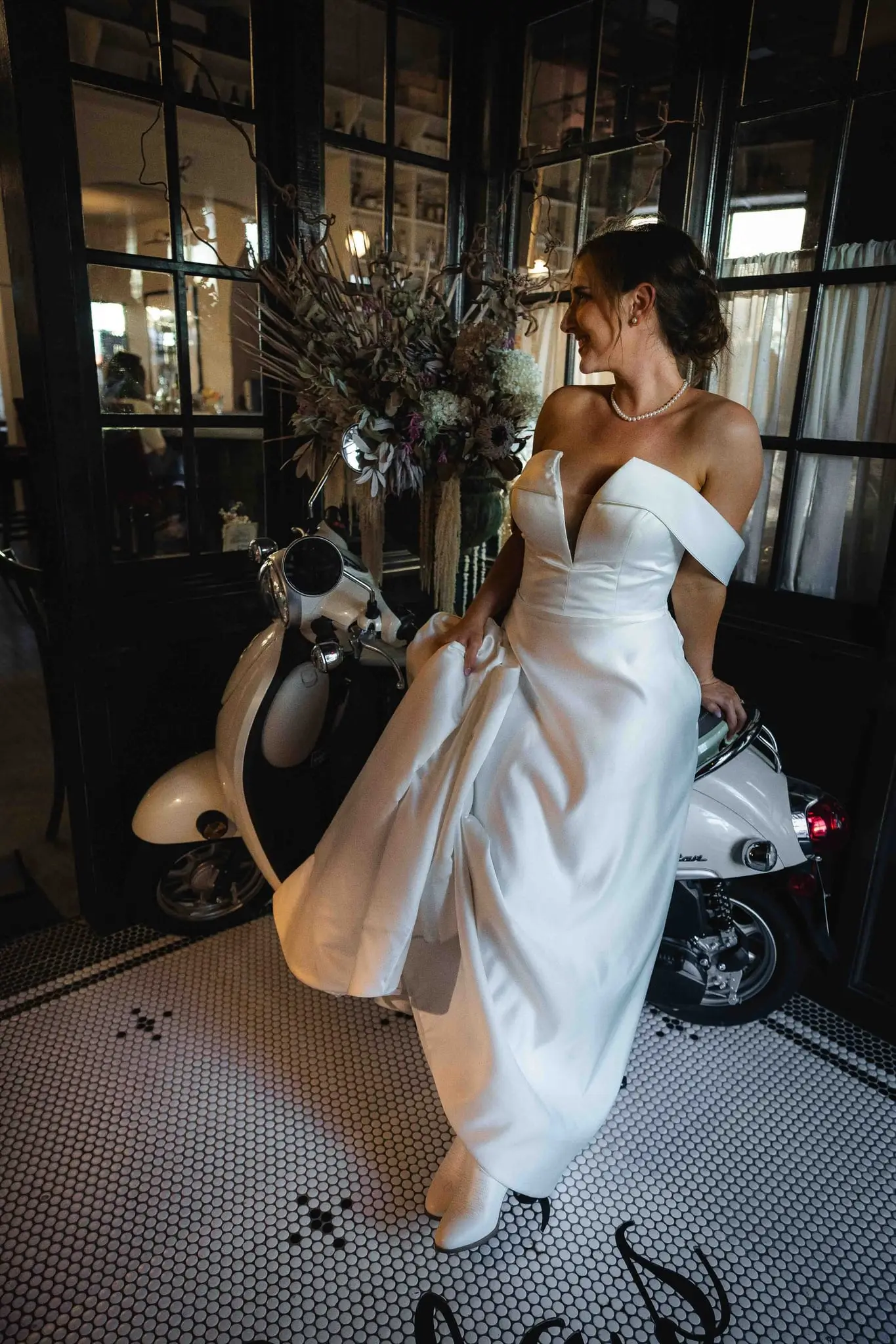 Bride sitting on a white vintage scooter in a tiled restaurant foyer, holding her gown