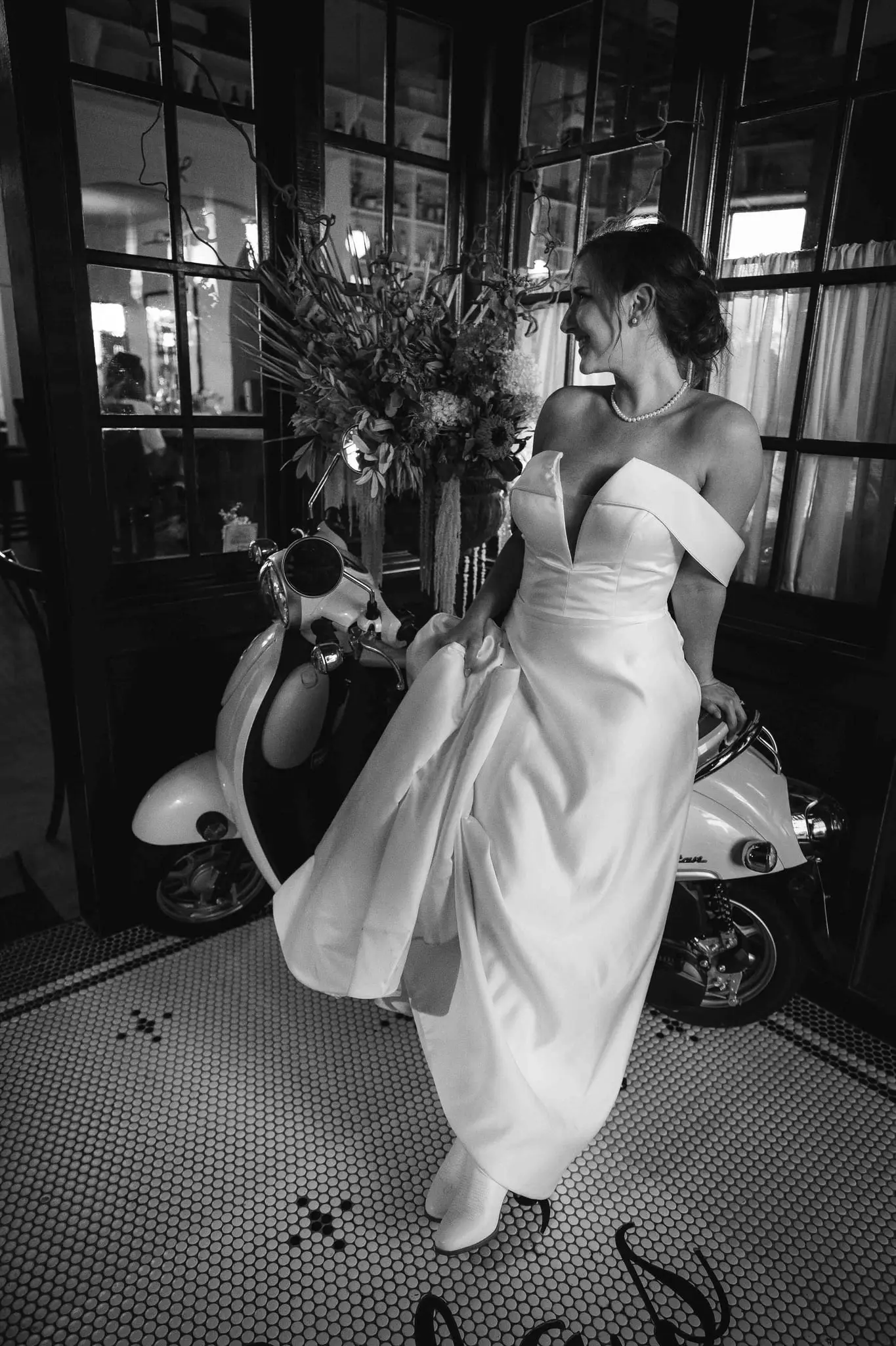 Black and white portrait of the bride perched on a vintage scooter beside a large floral arrangement