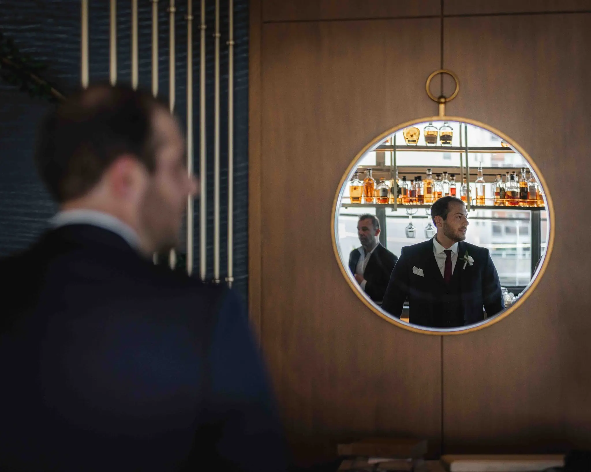 Groom in a navy suit reflected in a round brass mirror above a hotel bar