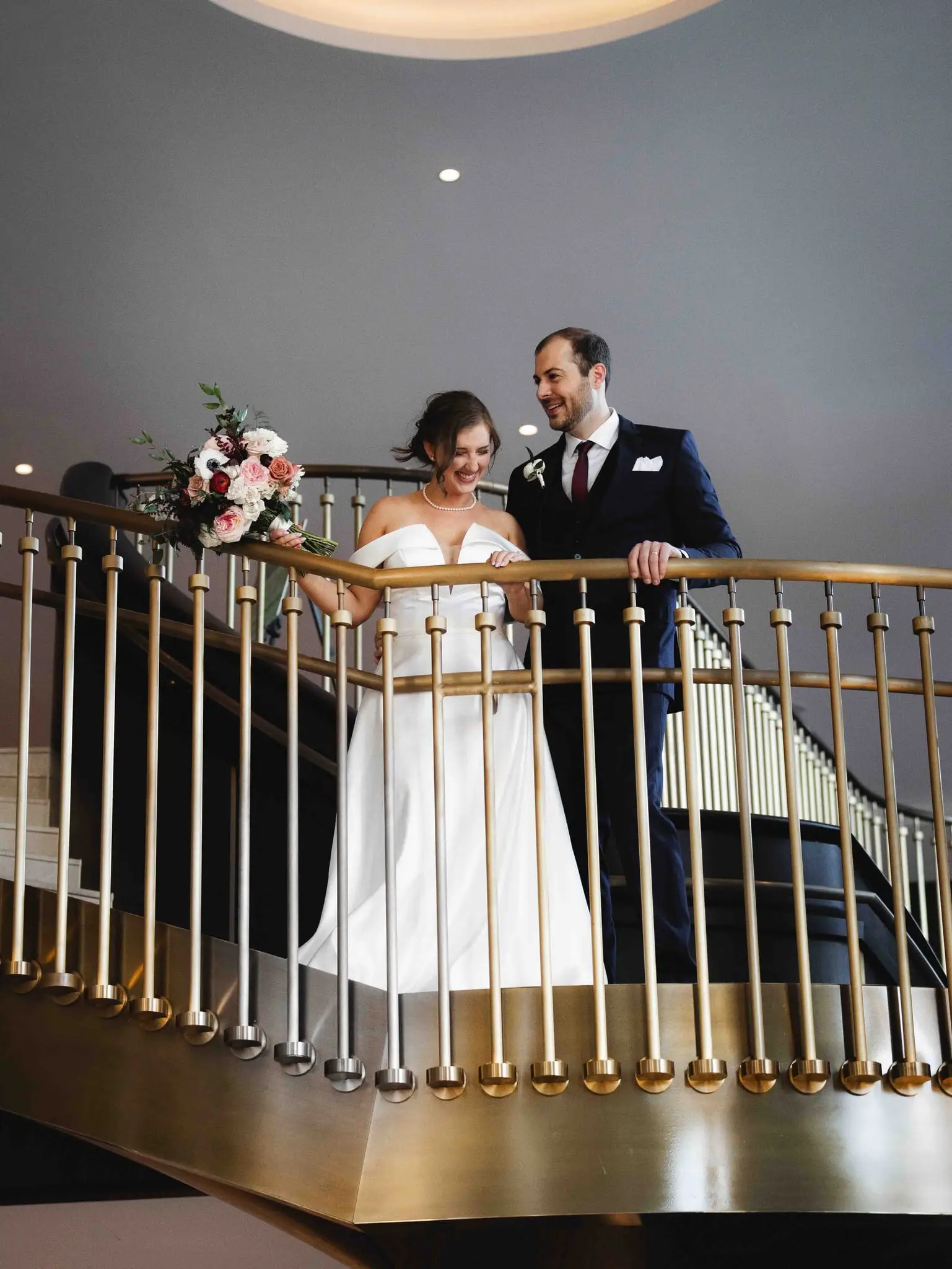 Ashley and Alex wedding at Conrad Nashville, Nashville TN — the couple on a brass-railed balcony above the lobby, the bride holding a bouquet