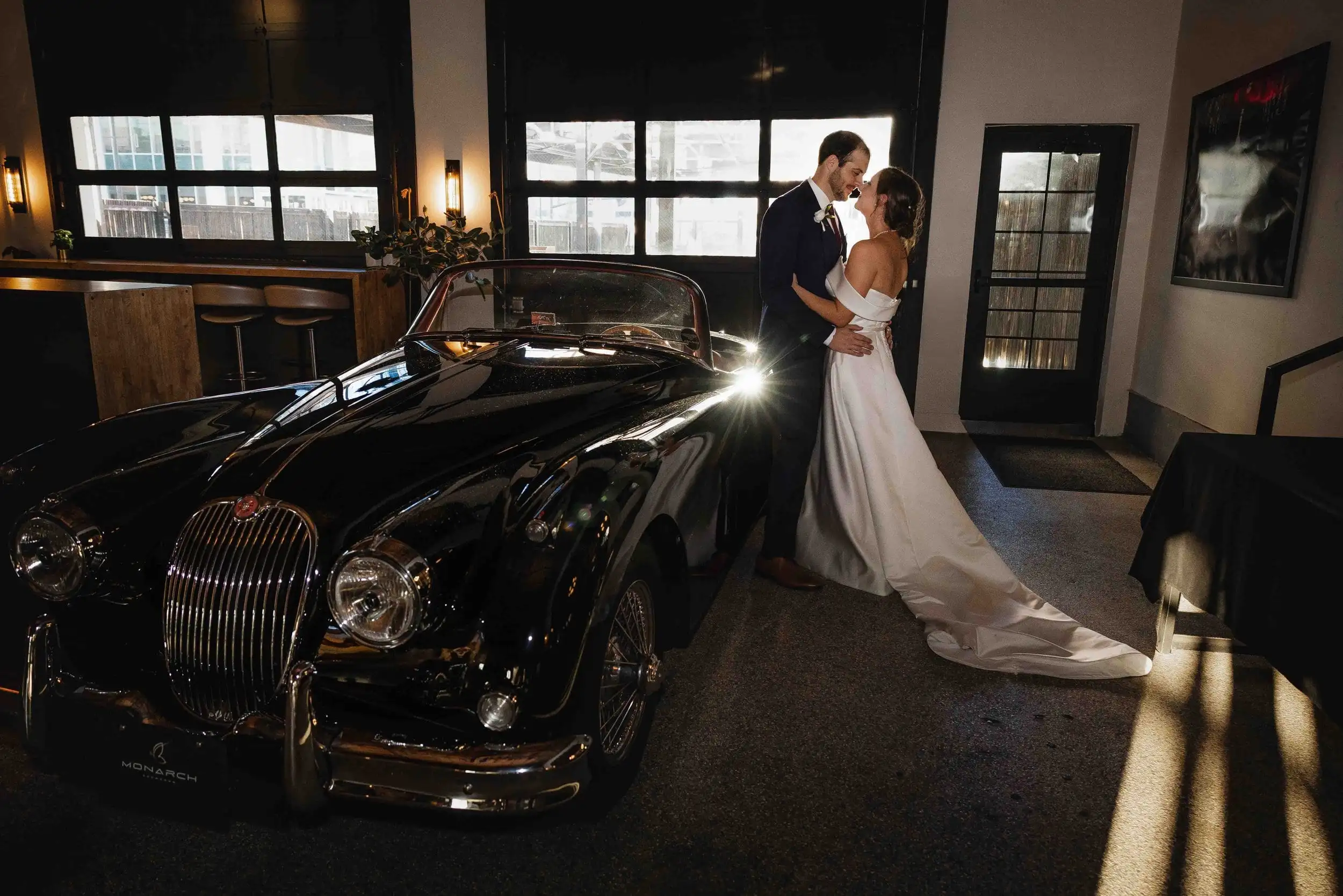 Couple about to kiss beside a vintage black convertible in a sunlit garage