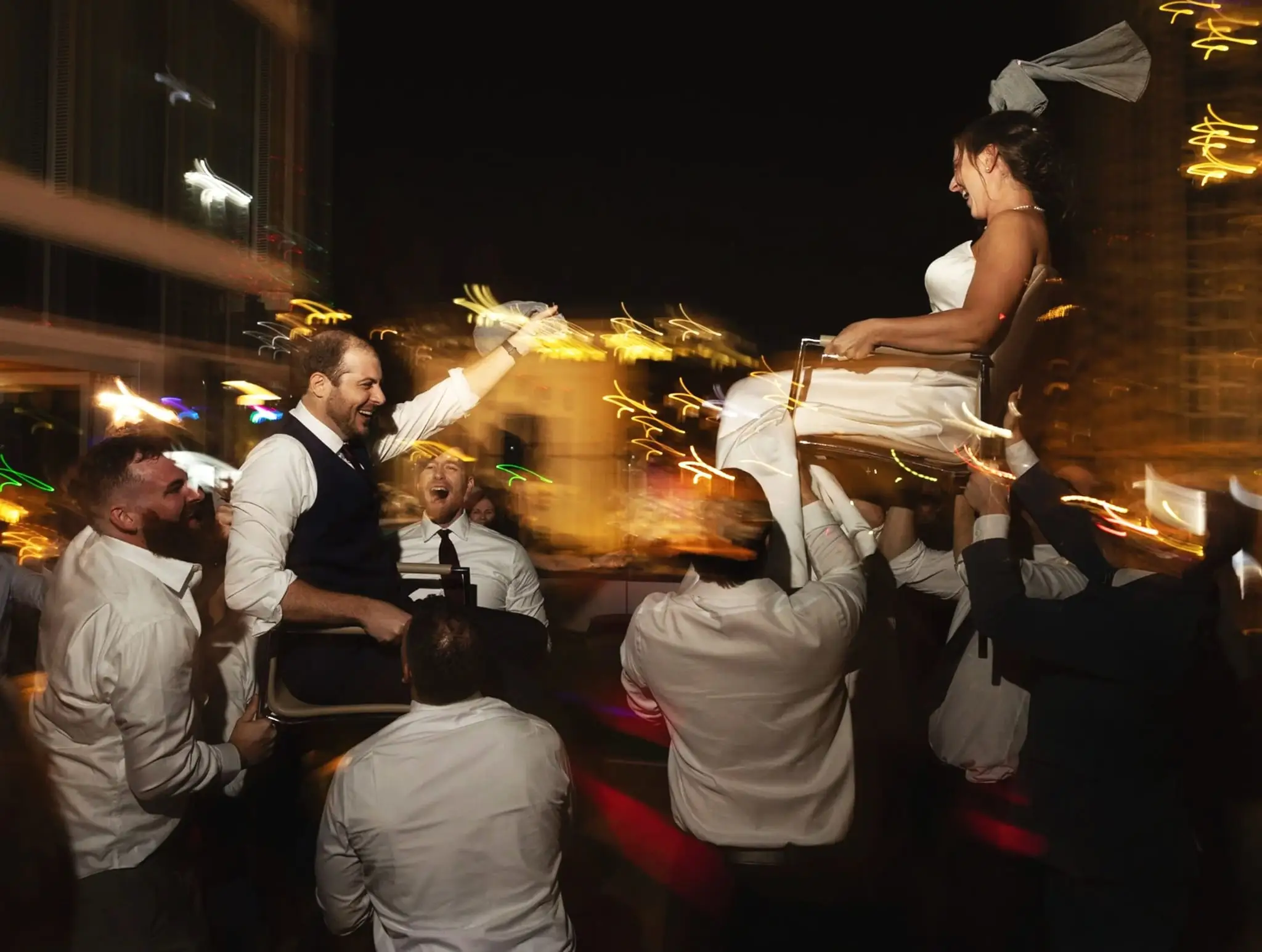 Guests lifting the bride in a chair over the dance floor at night, city lights streaking behind