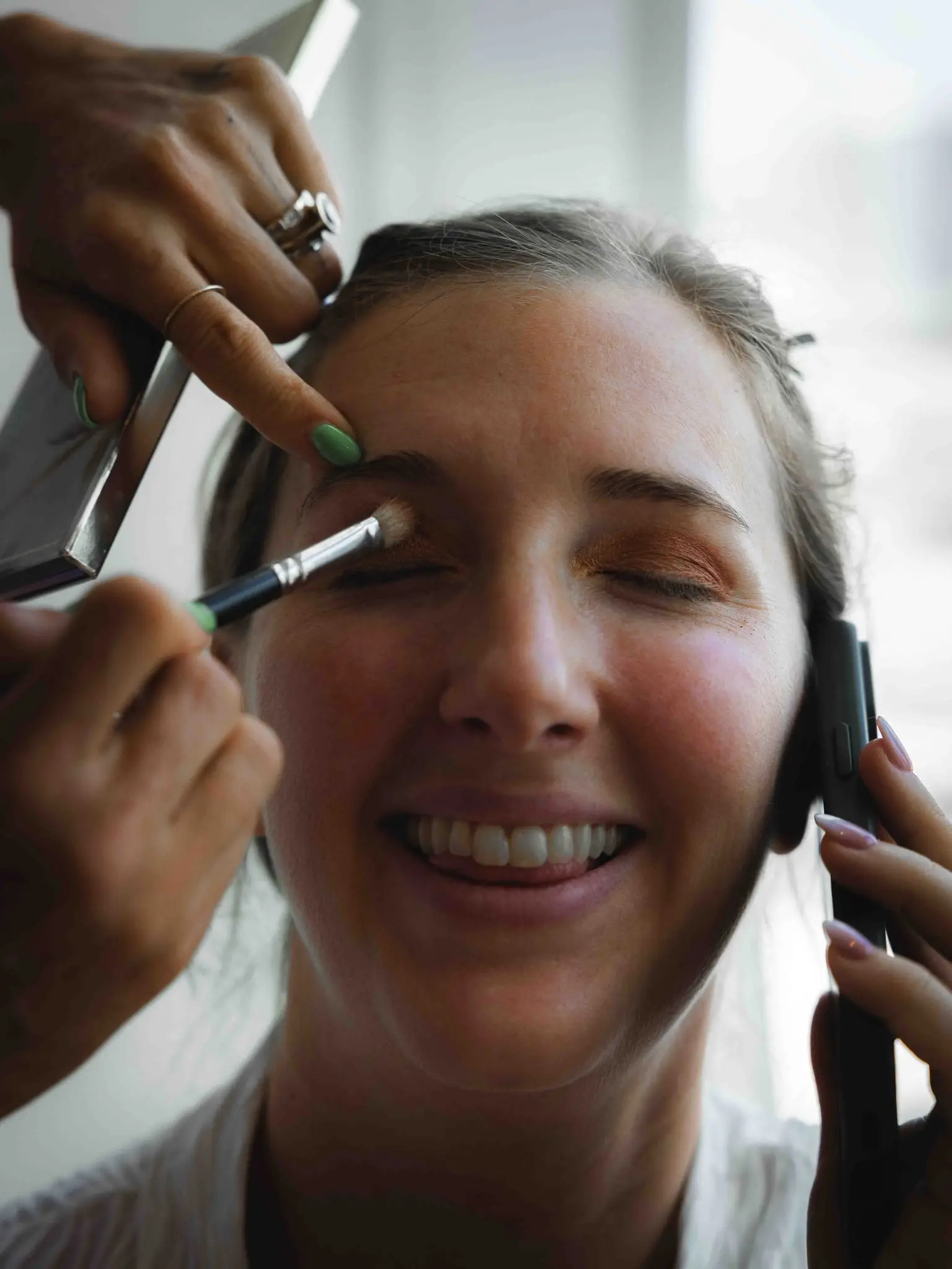 Bride laughing on the phone while a makeup artist applies her eyeshadow