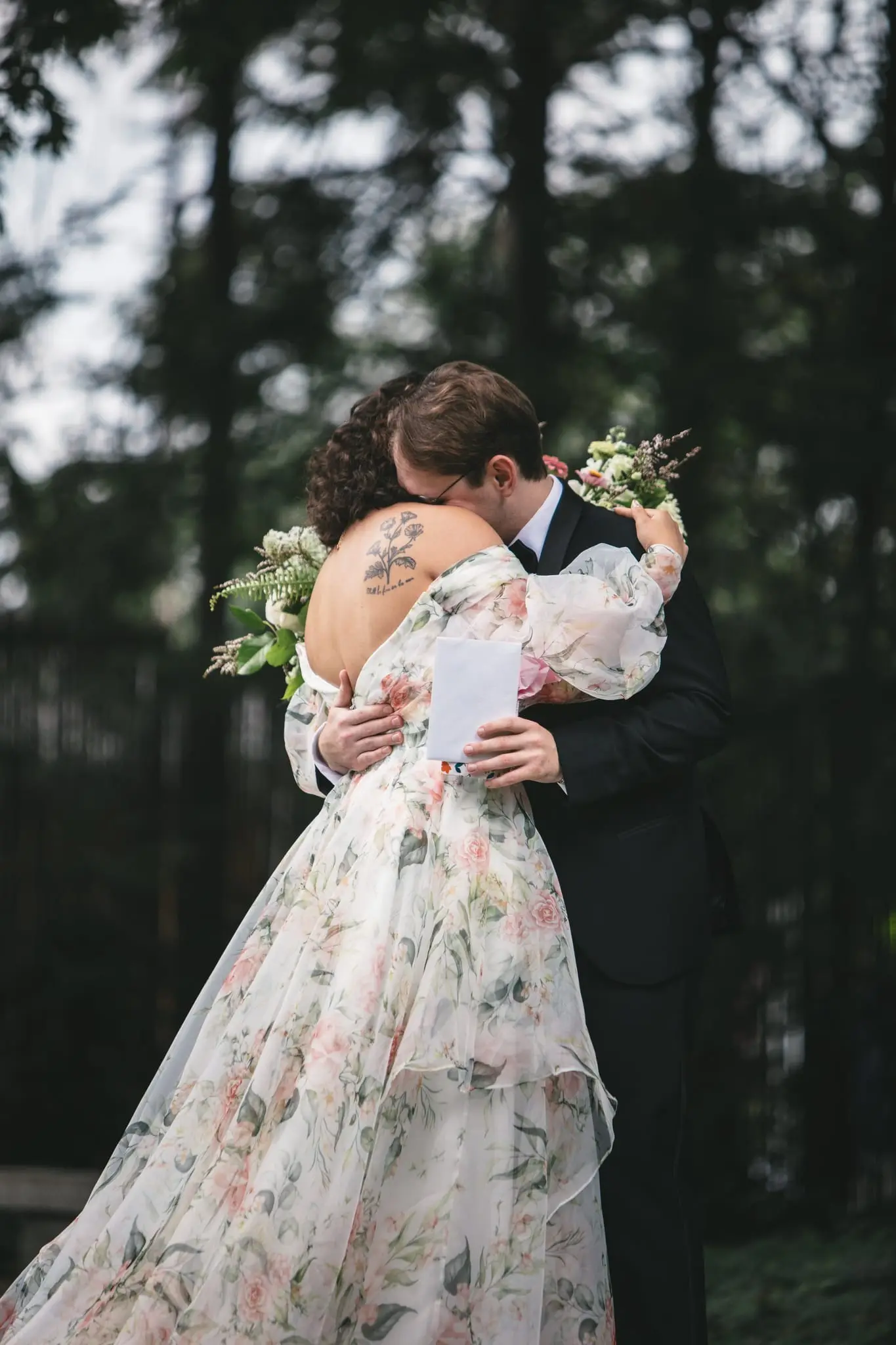 Bride and groom hugging in the pines, her bouquet and a folded note in hand
