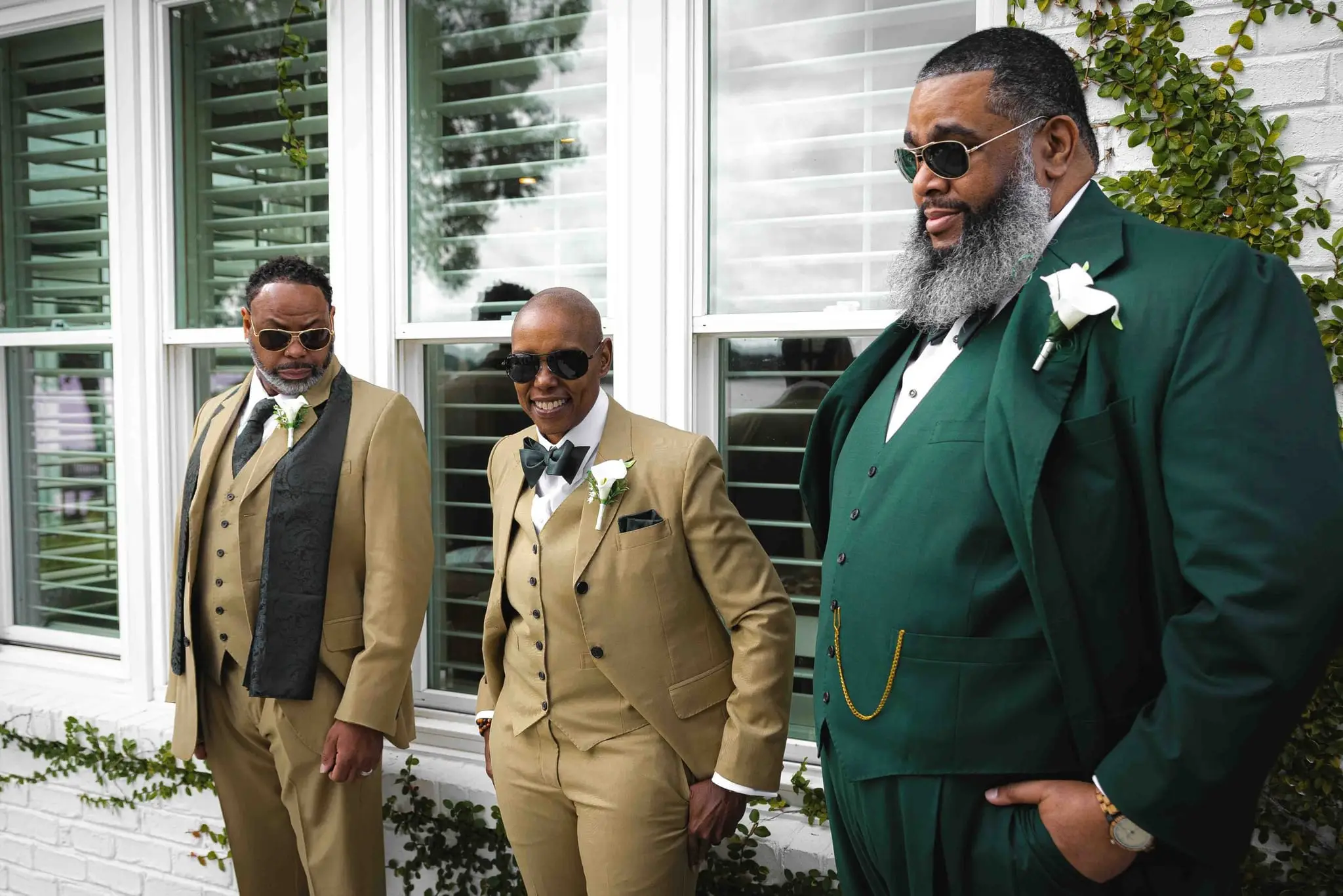 Groom in a green suit with two groomsmen in tan suits and sunglasses by a white house