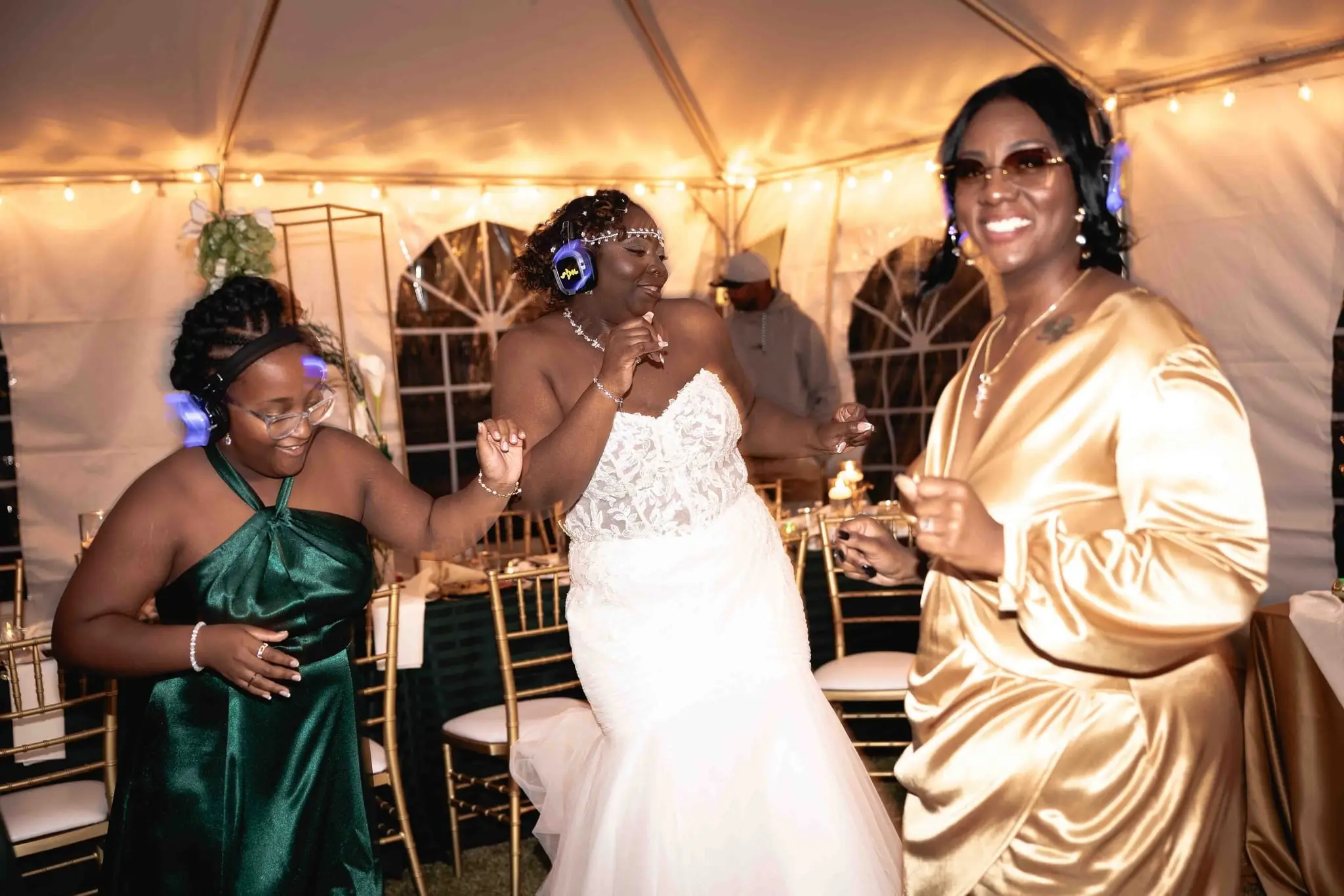 Bride dancing with a bridesmaid and a guest in gold satin, all wearing glowing silent disco headphones
