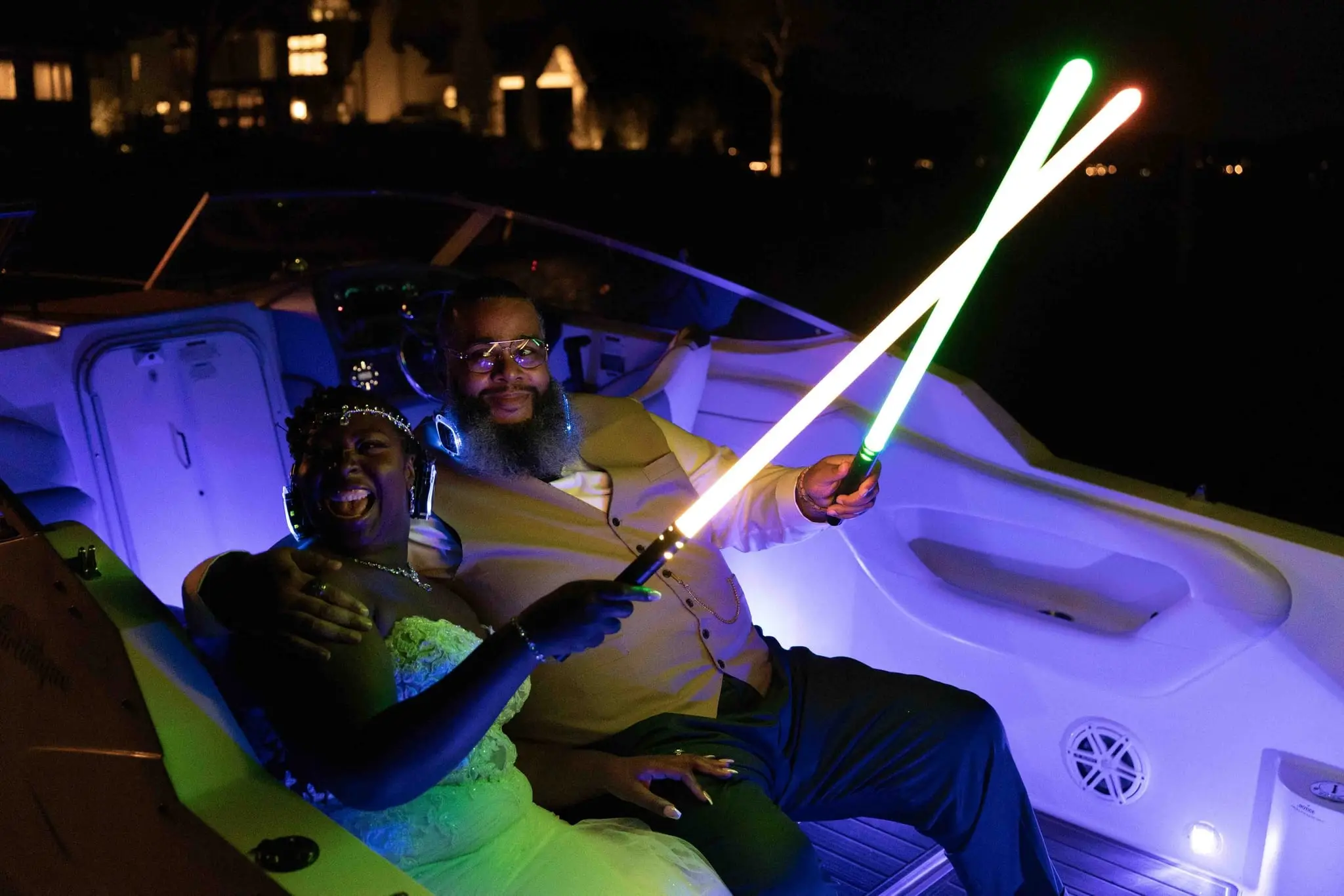 Bride and groom lounging on a blue-lit boat at night, crossing glowing light sticks