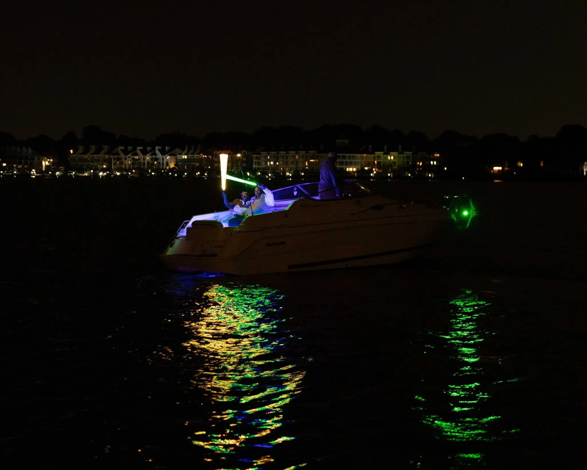 Boat glowing with blue lights on the dark lake at night, green reflections on the water and a lit shoreline