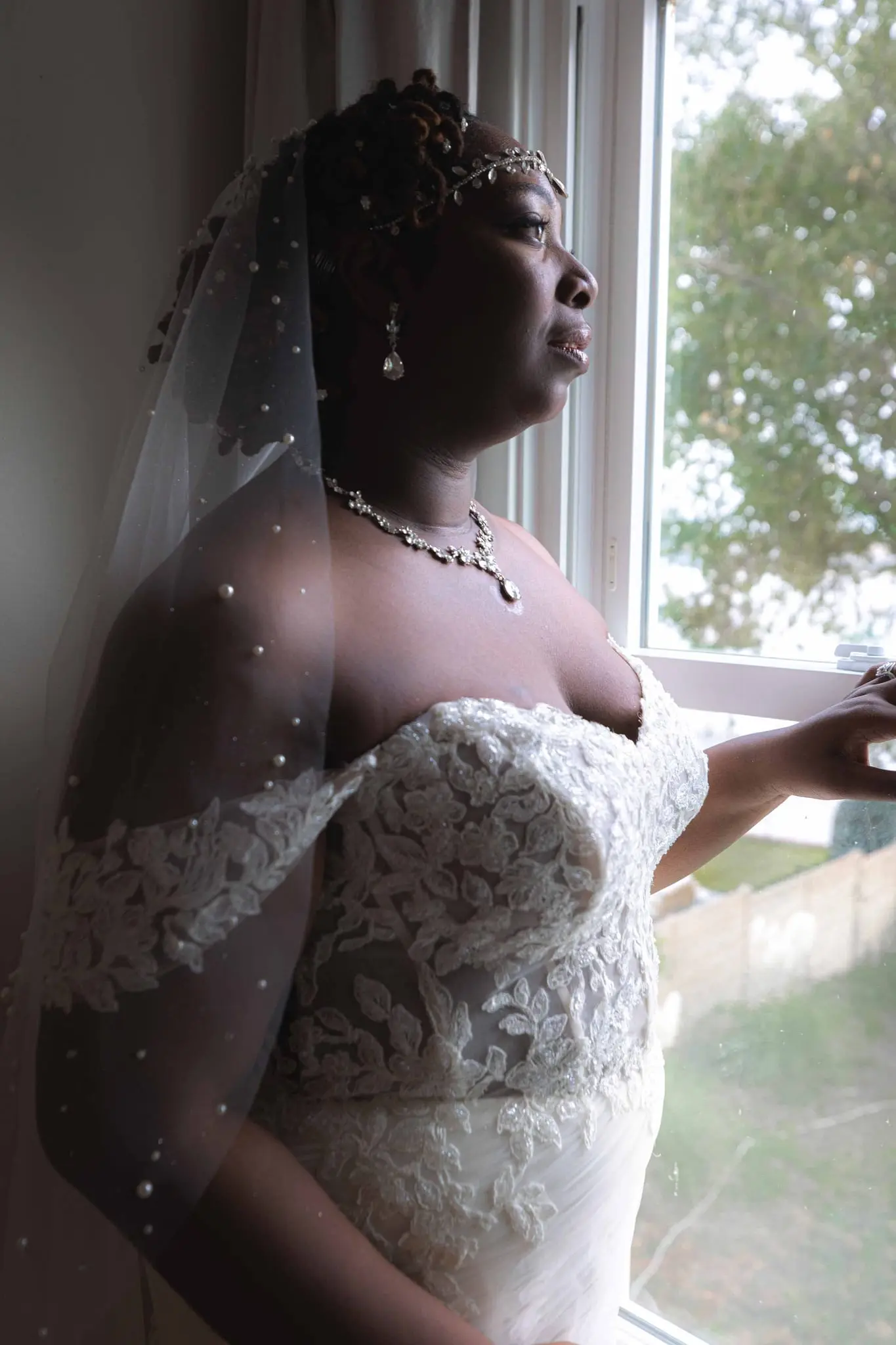 Bride in a lace off-shoulder gown and beaded veil gazing out a window