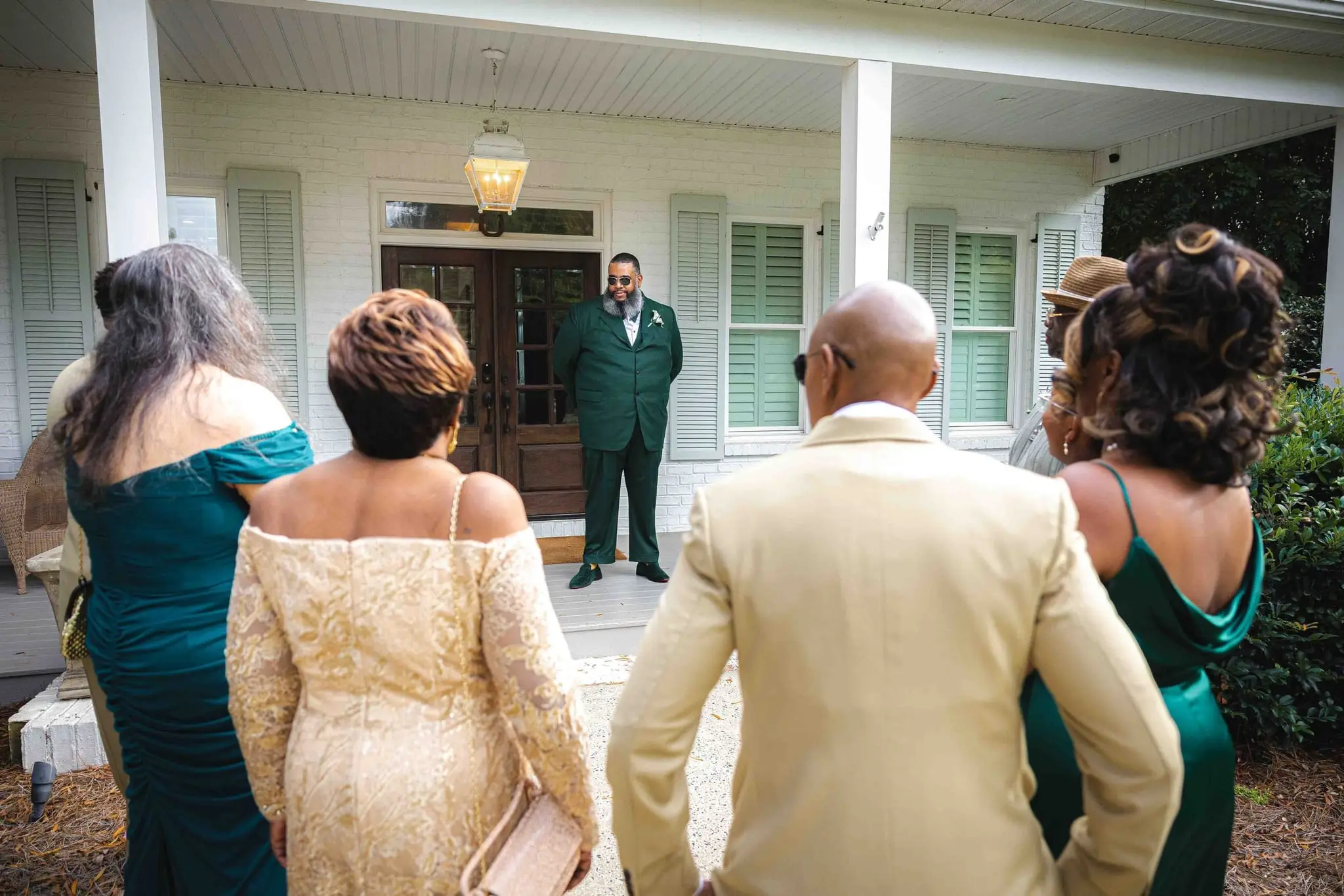 Family and guests gather facing a porch where the groom waits in a green suit