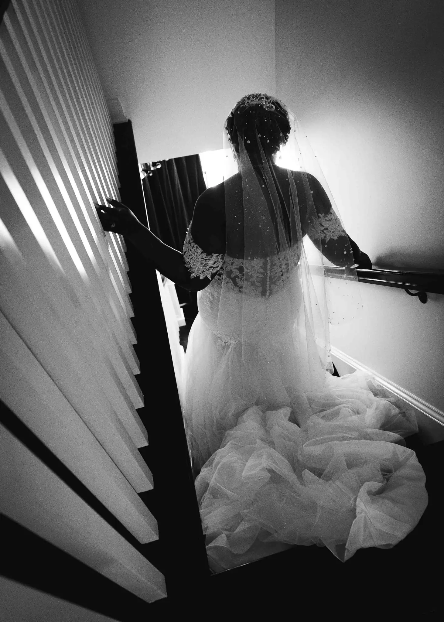Black and white view of the bride from behind, veil trailing, on a staircase