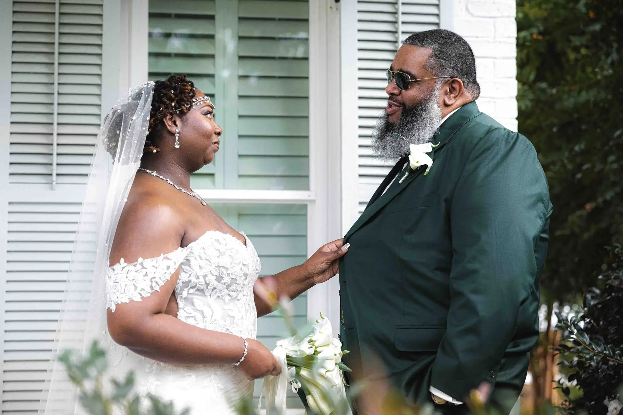 Bride gently straightening the groom's lapel as they face each other beside green shutters