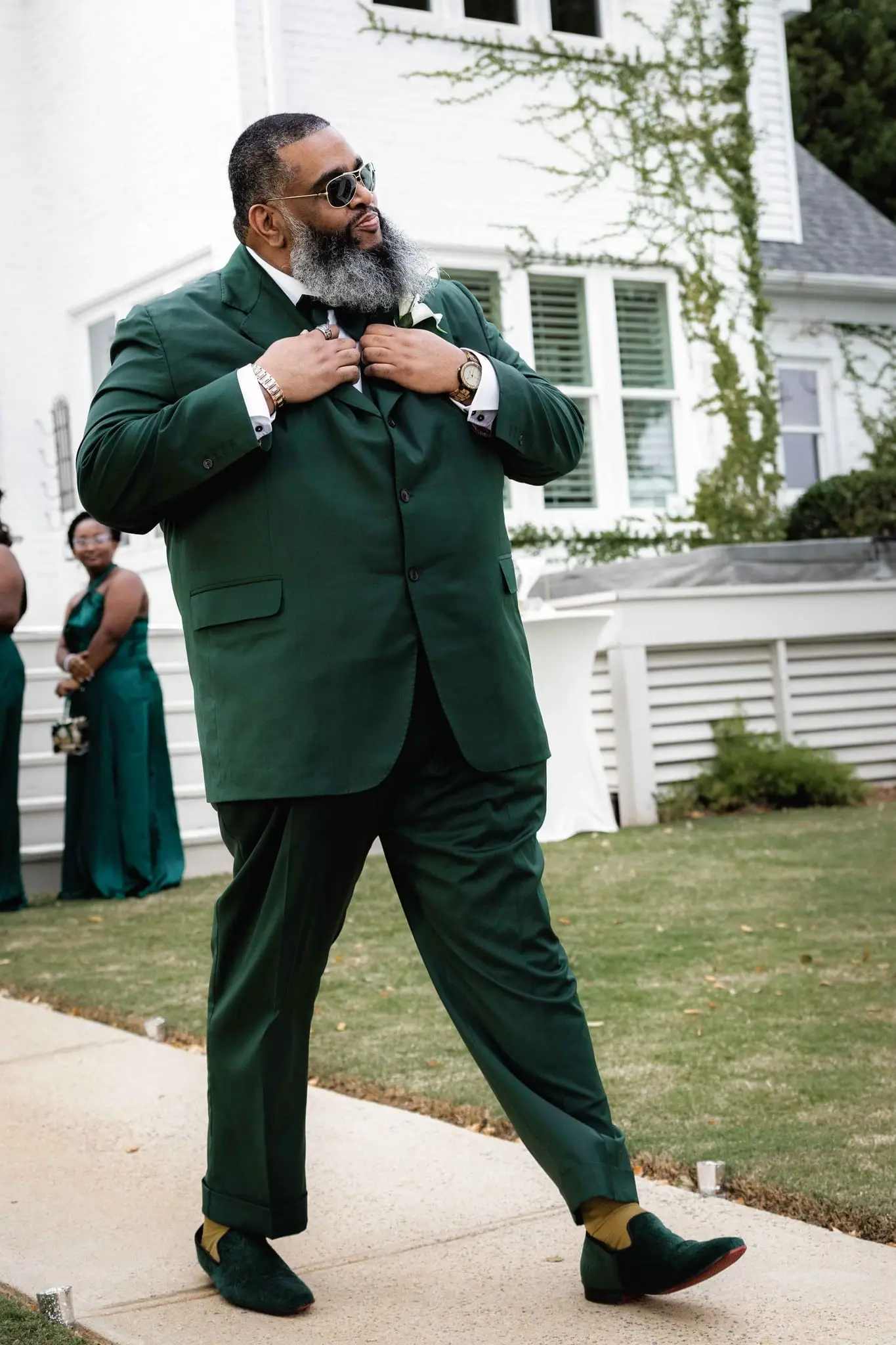 Groom in an emerald suit and sunglasses strutting up the sidewalk and adjusting his bow tie