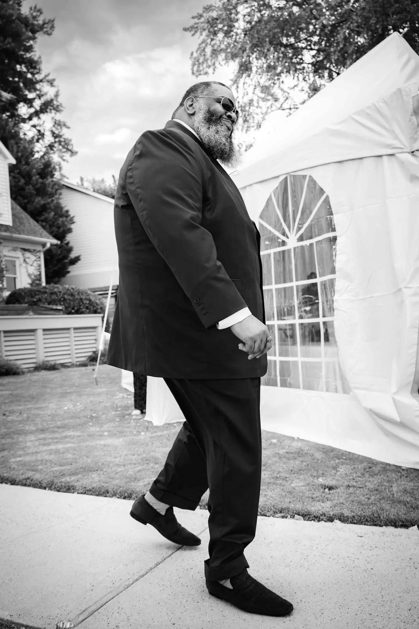 Black and white portrait of the groom in a dark suit striding past a white event tent