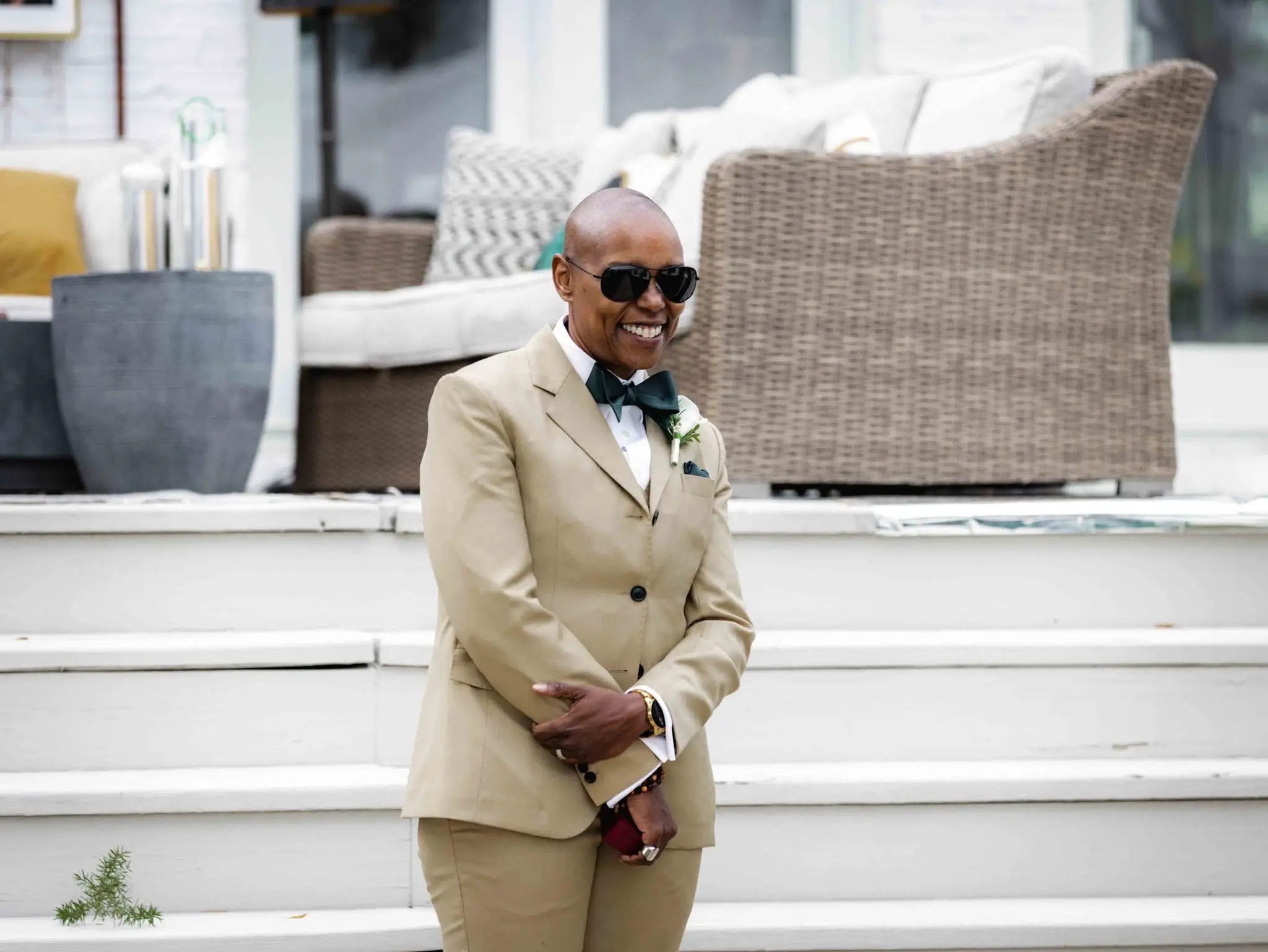Groomsman in a tan suit and bow tie smiling on white steps beside a wicker sofa