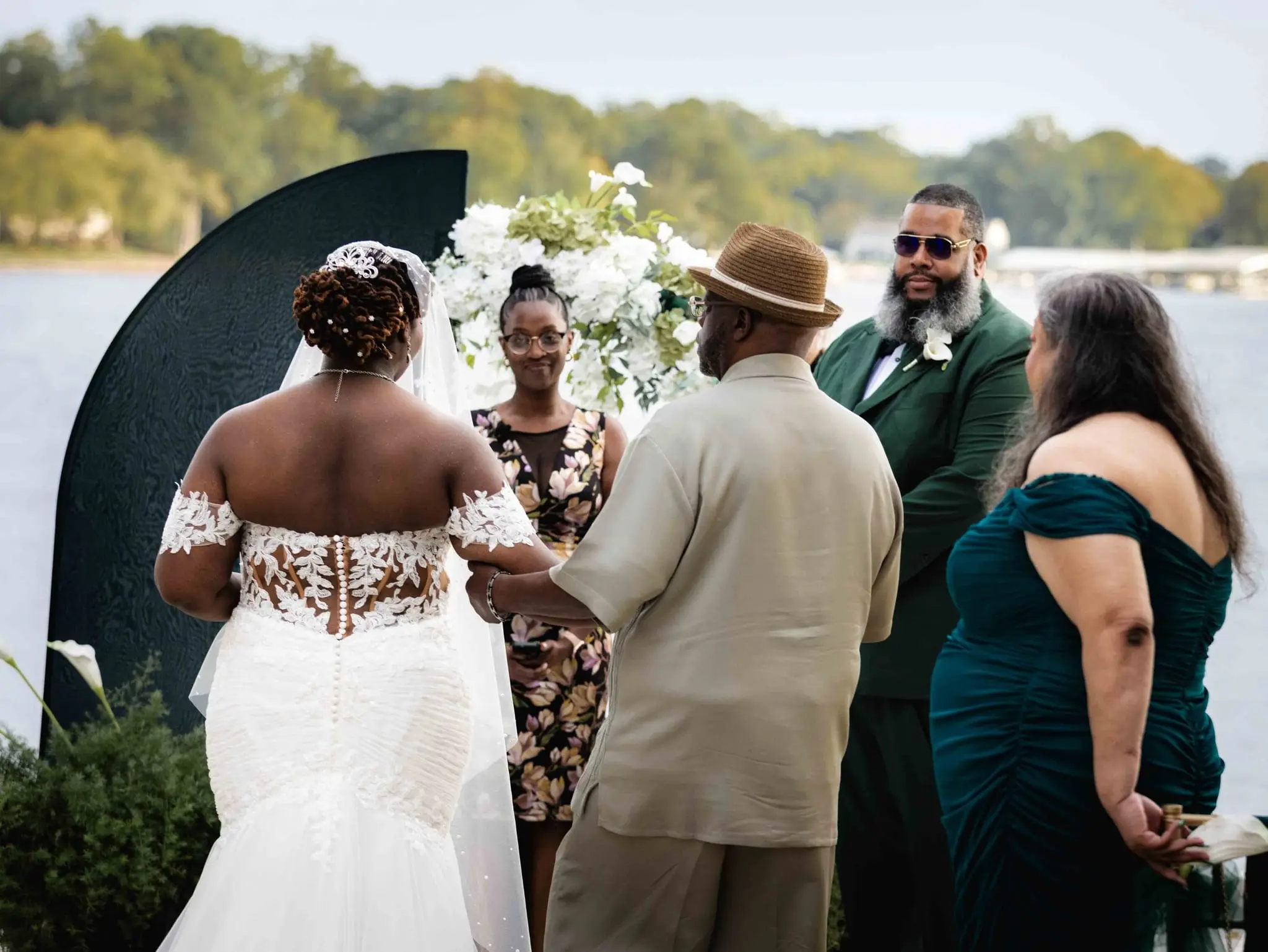 Bride at the ceremony arch as an escort in a tan suit presents her to the groom by the lake