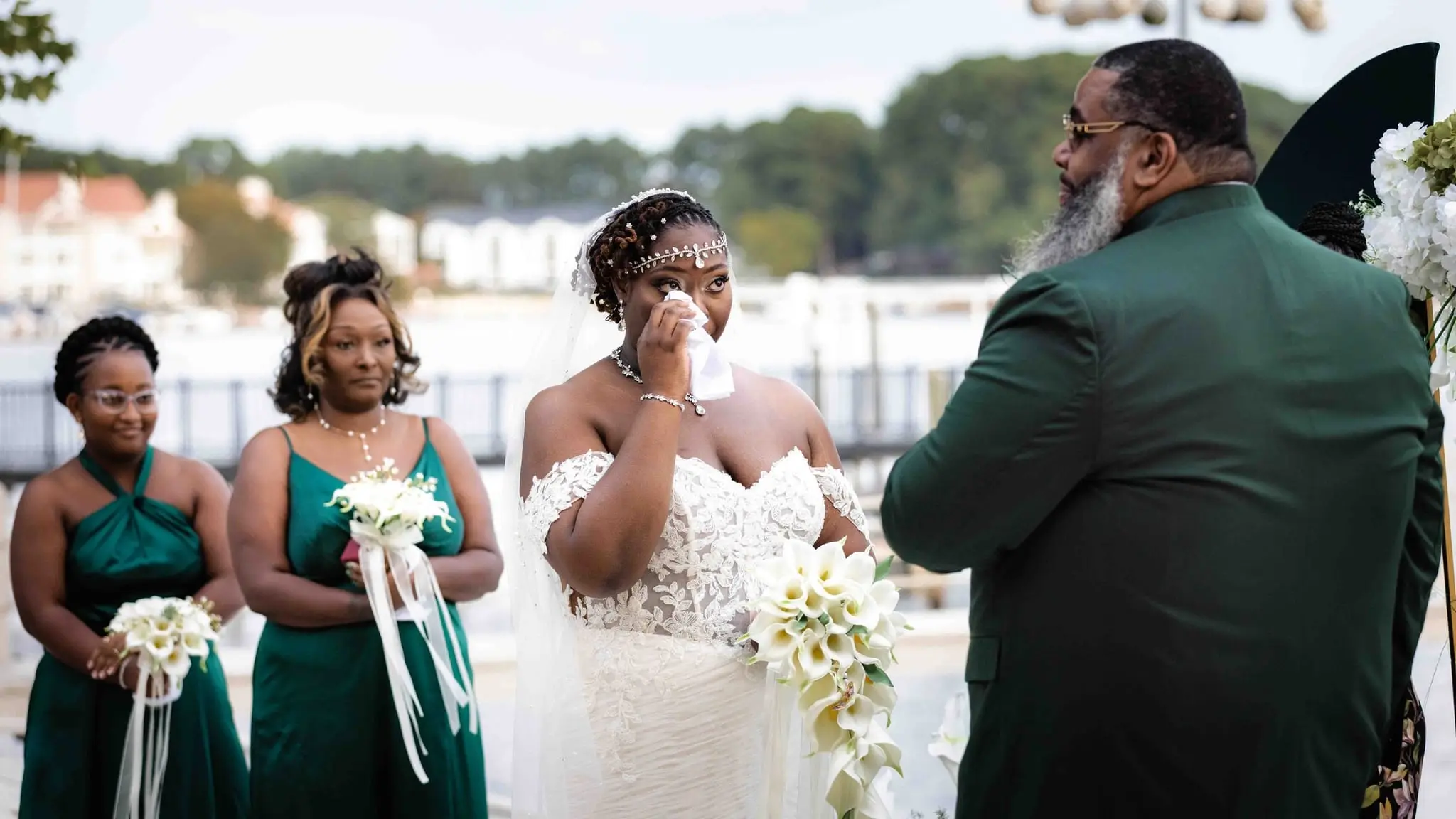 Bride dabbing a tear as bridesmaids in green hold bouquets beside the groom