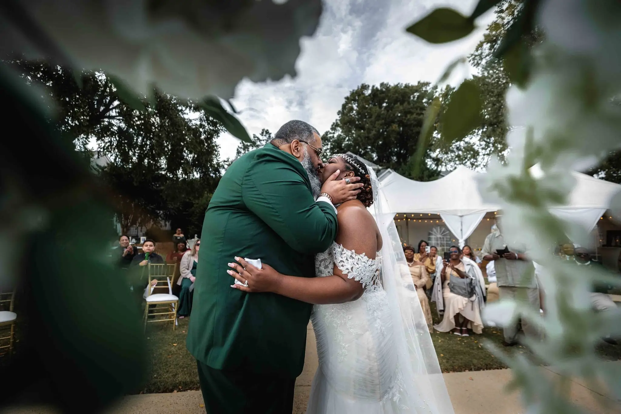 Groom and bride embracing at the end of the aisle, framed by leaves, guests under a tent behind
