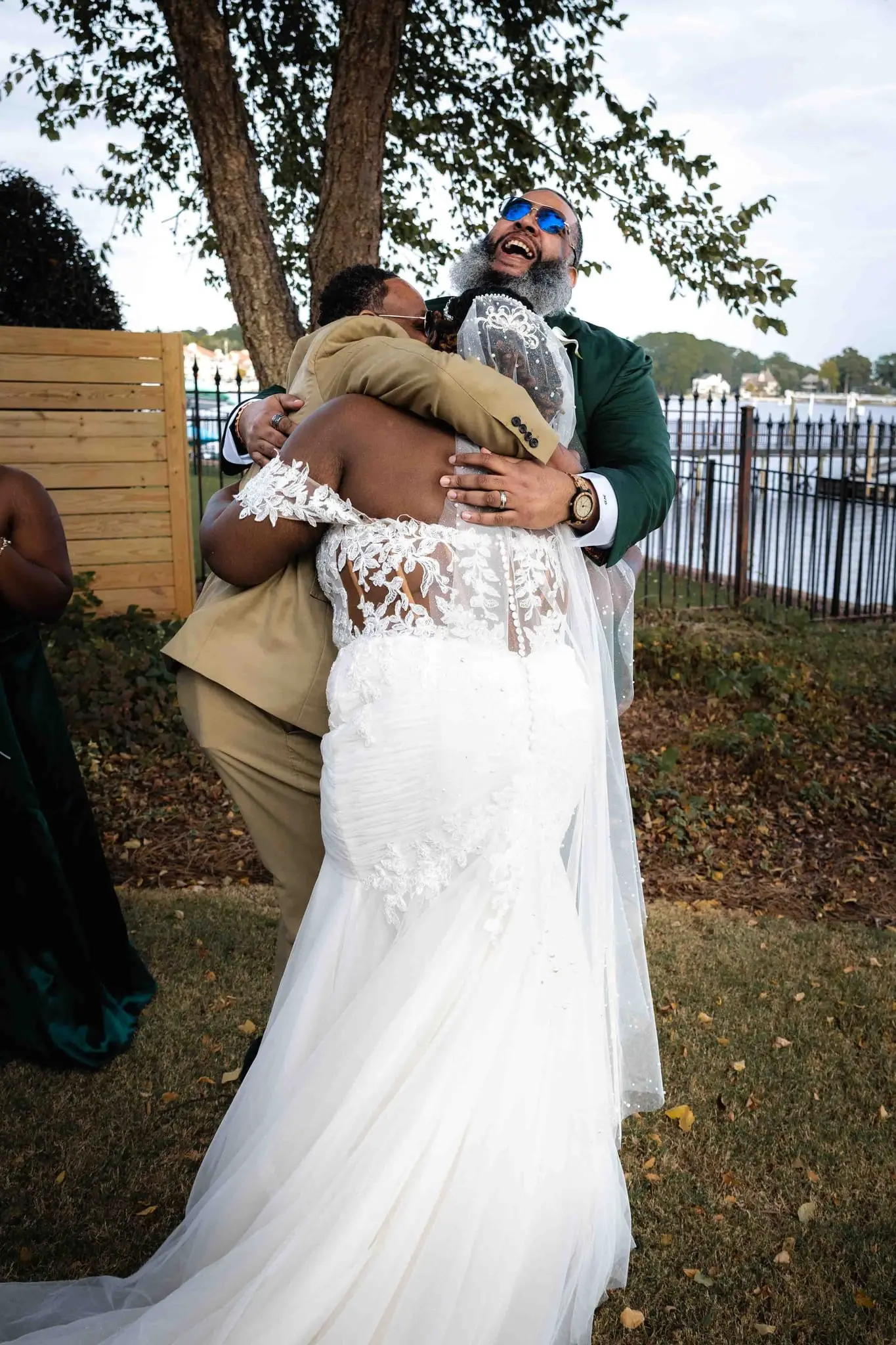 Bride in a lace-back gown hugging a guest in a tan suit as the groom laughs behind them