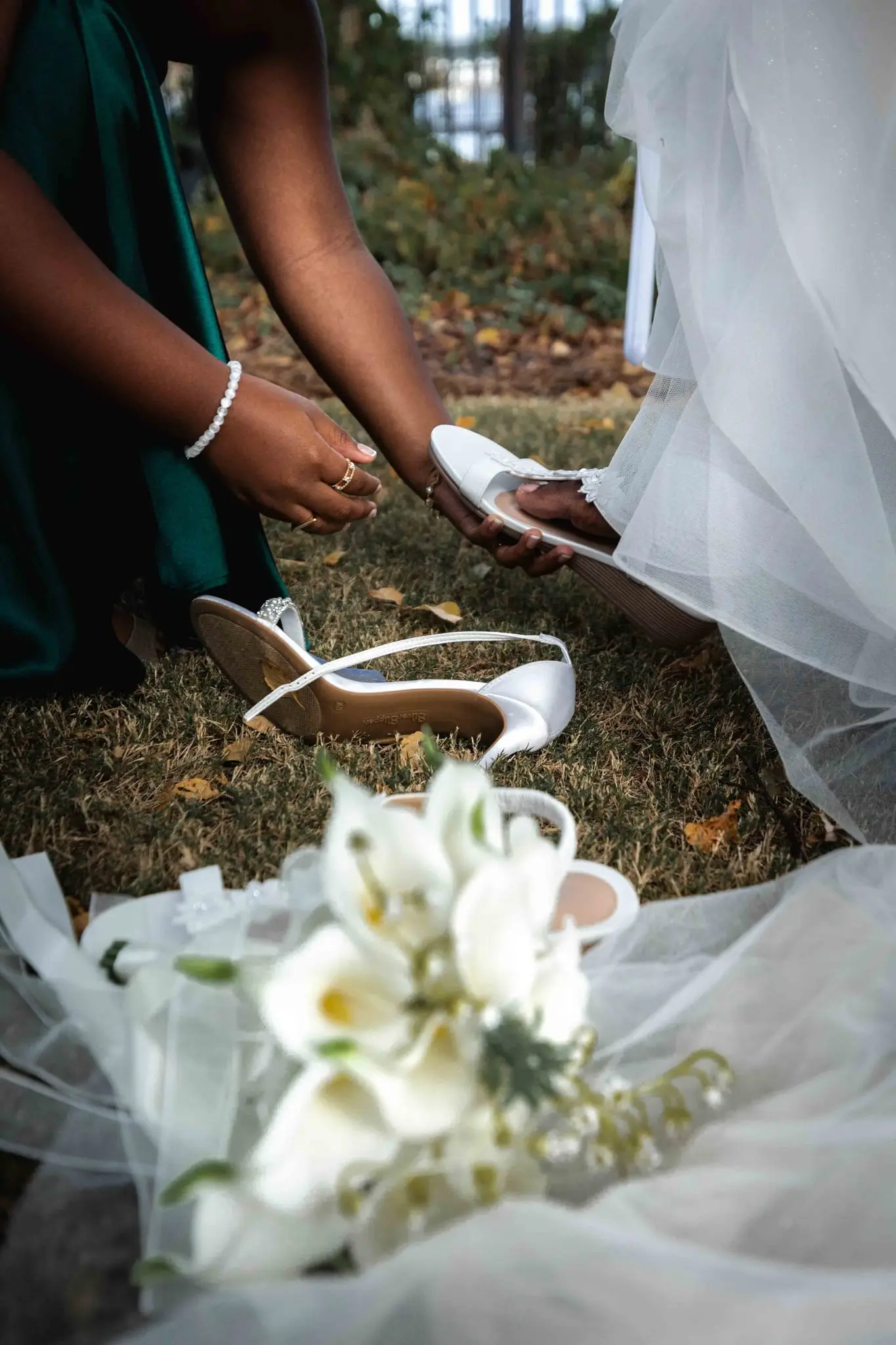 Bridesmaid in emerald satin helping the bride into white heels beside her calla lily bouquet