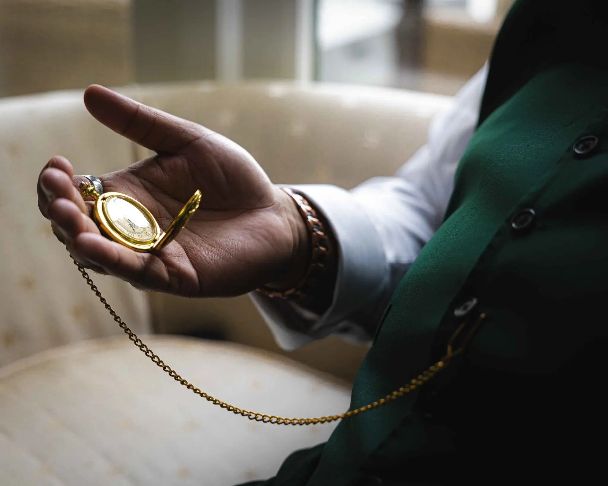 Groom's hand holding an open gold pocket watch on a chain, his green vest sleeve beside it