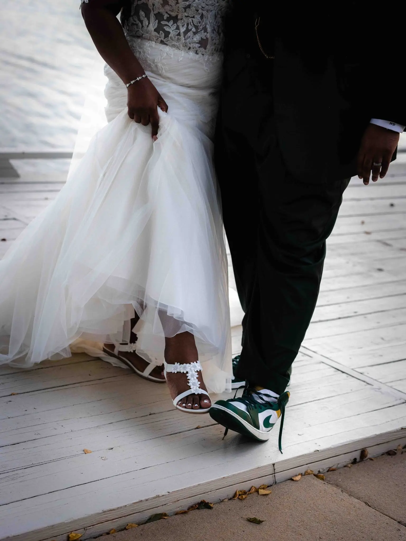 Close-up of the bride's lifted gown and white lace sandals beside the groom's green and white sneakers